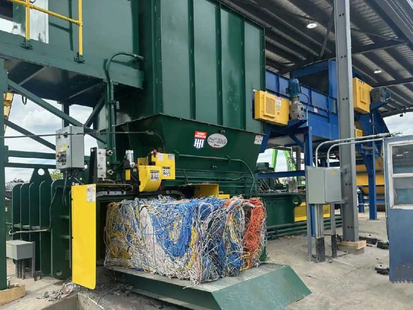 Industrial baler compressing colorful wires into a dense bale at a recycling facility under a metal roof.