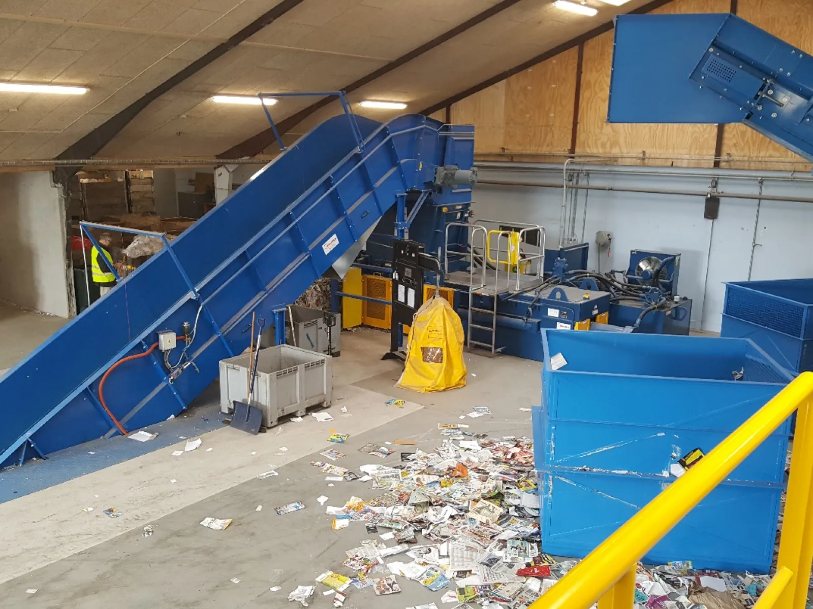 Recycling plant interior with blue conveyor belts, yellow railings, and scattered paper waste on concrete floor.