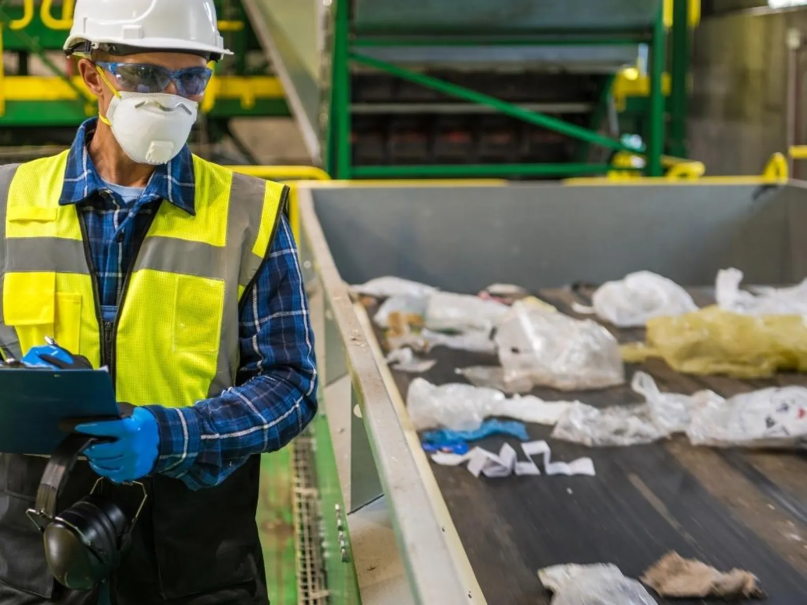 Worker wearing safety gear inspects plastic waste on conveyor belt at recycling facility.