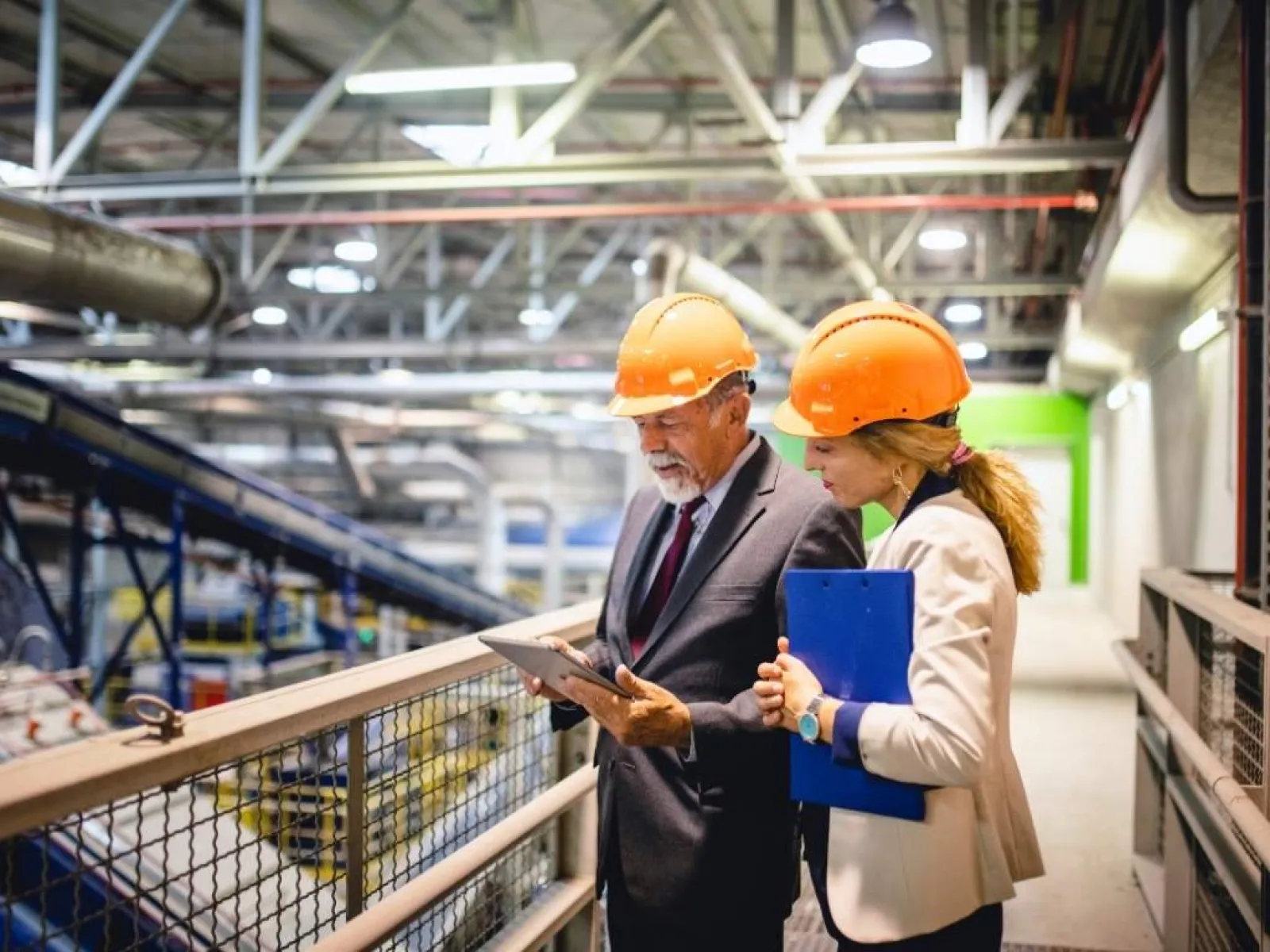 Two professionals wearing orange safety helmets inspecting factory operations using a tablet and clipboard.