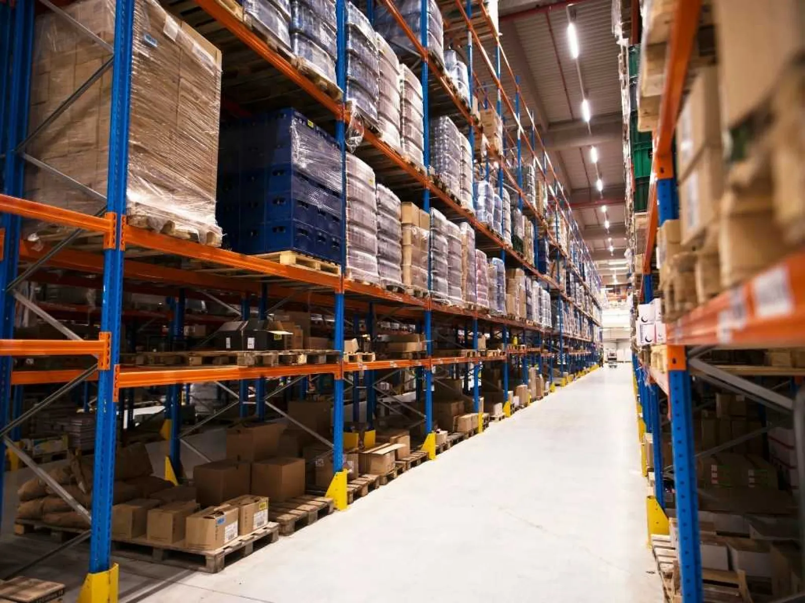 Warehouse aisle with tall metal racks filled with pallets of boxes and goods under bright industrial lighting