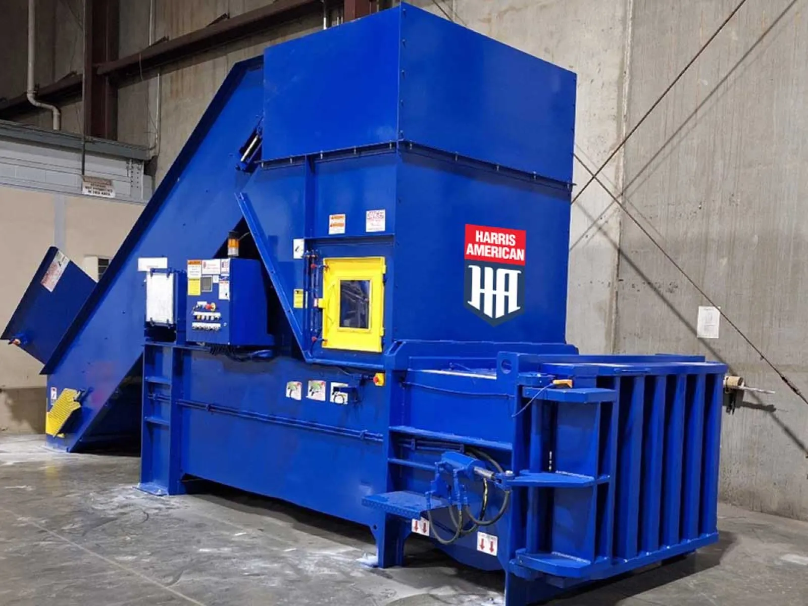 Large blue industrial Harris American baler machine in a warehouse with concrete floors and walls.