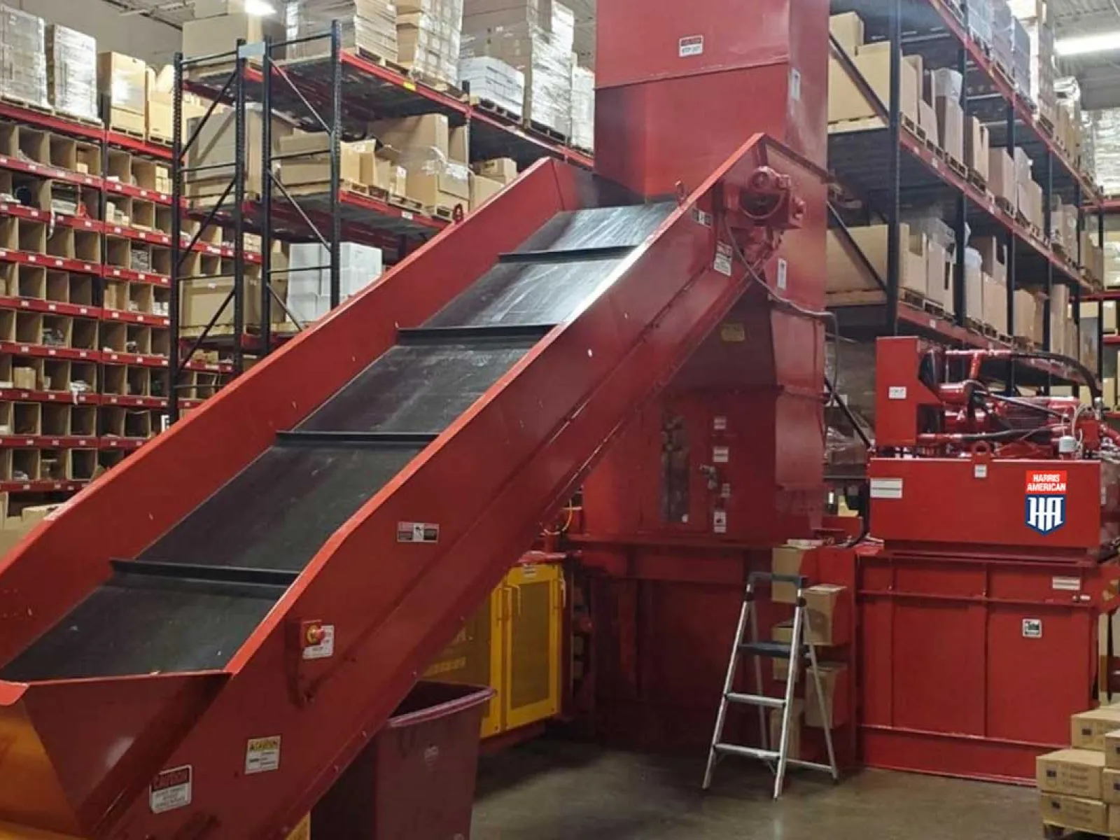 Red industrial conveyor belt system inside a warehouse with shelves full of boxes on both sides.
