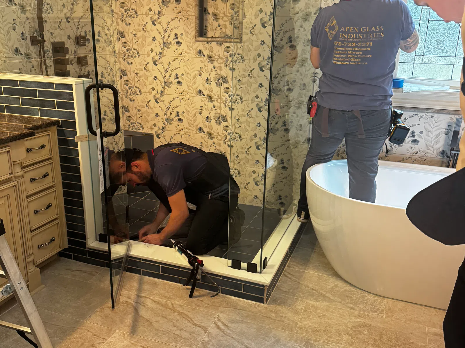 Two workers installing a glass shower enclosure in a floral tiled bathroom with modern fixtures and a freestanding tub.