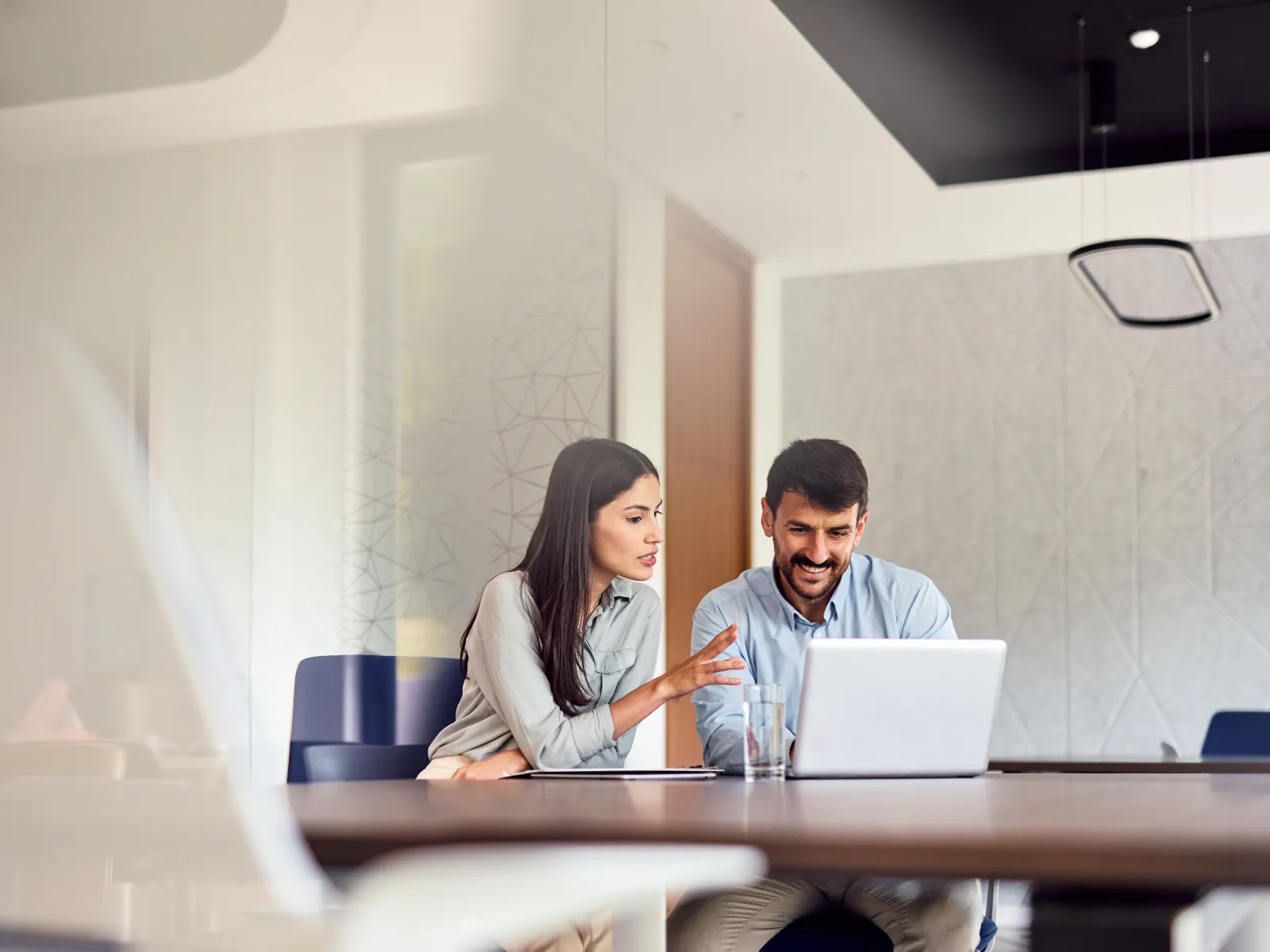 Two colleagues discussing work and looking at a laptop screen in a modern office meeting room.
