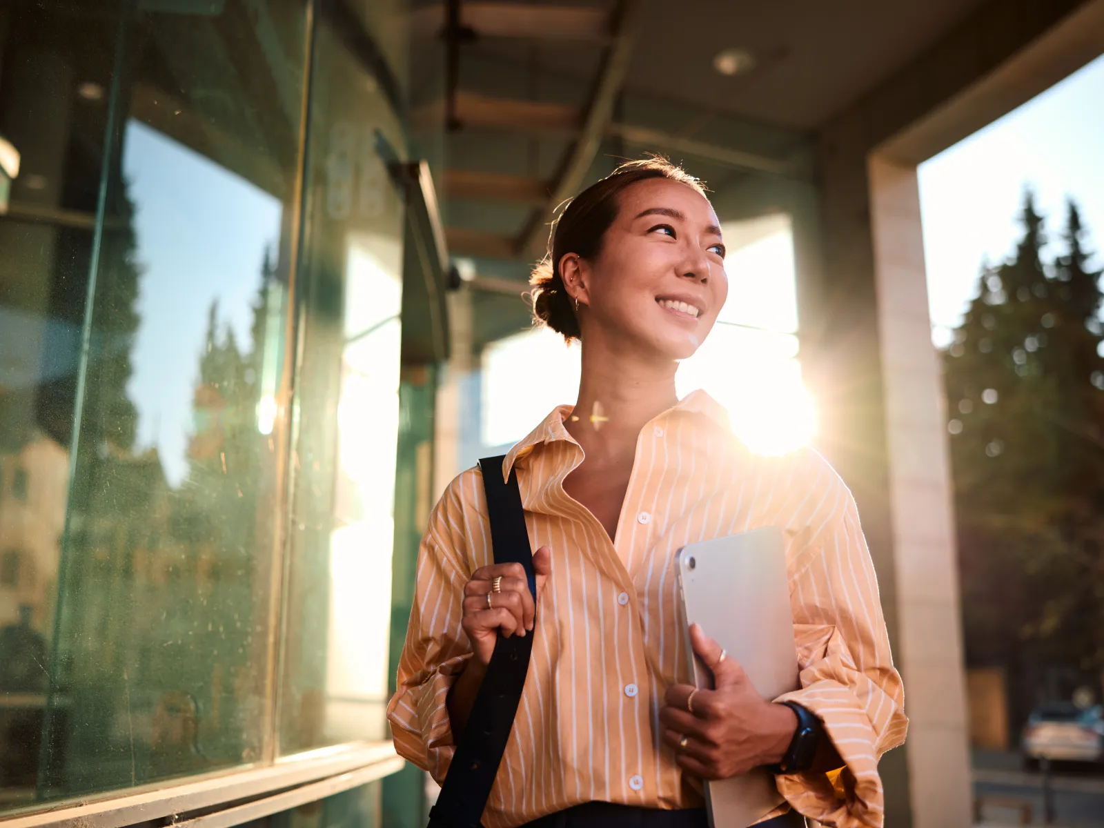 Young woman in striped shirt holding laptop and backpack, smiling outdoors with sunlight and glass reflections.