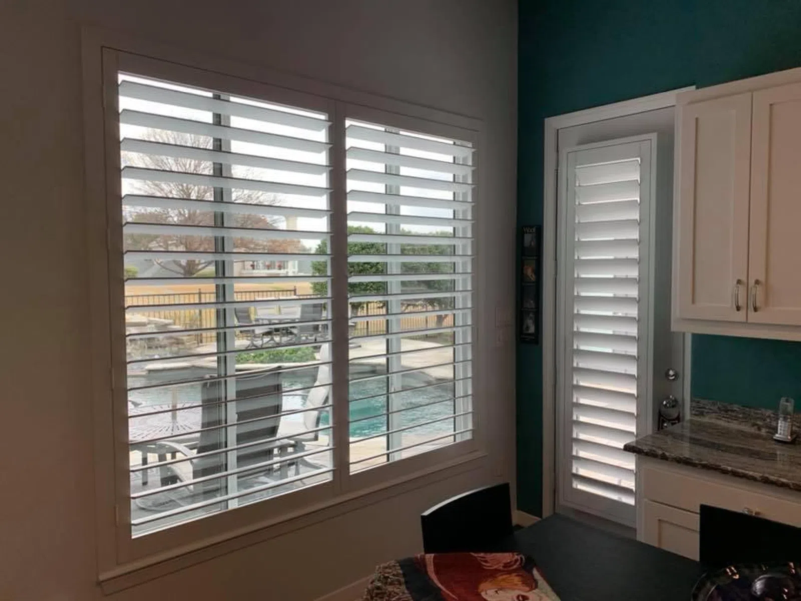 Kitchen view with white plantation shutters on windows and door overlooking a backyard pool and patio area.