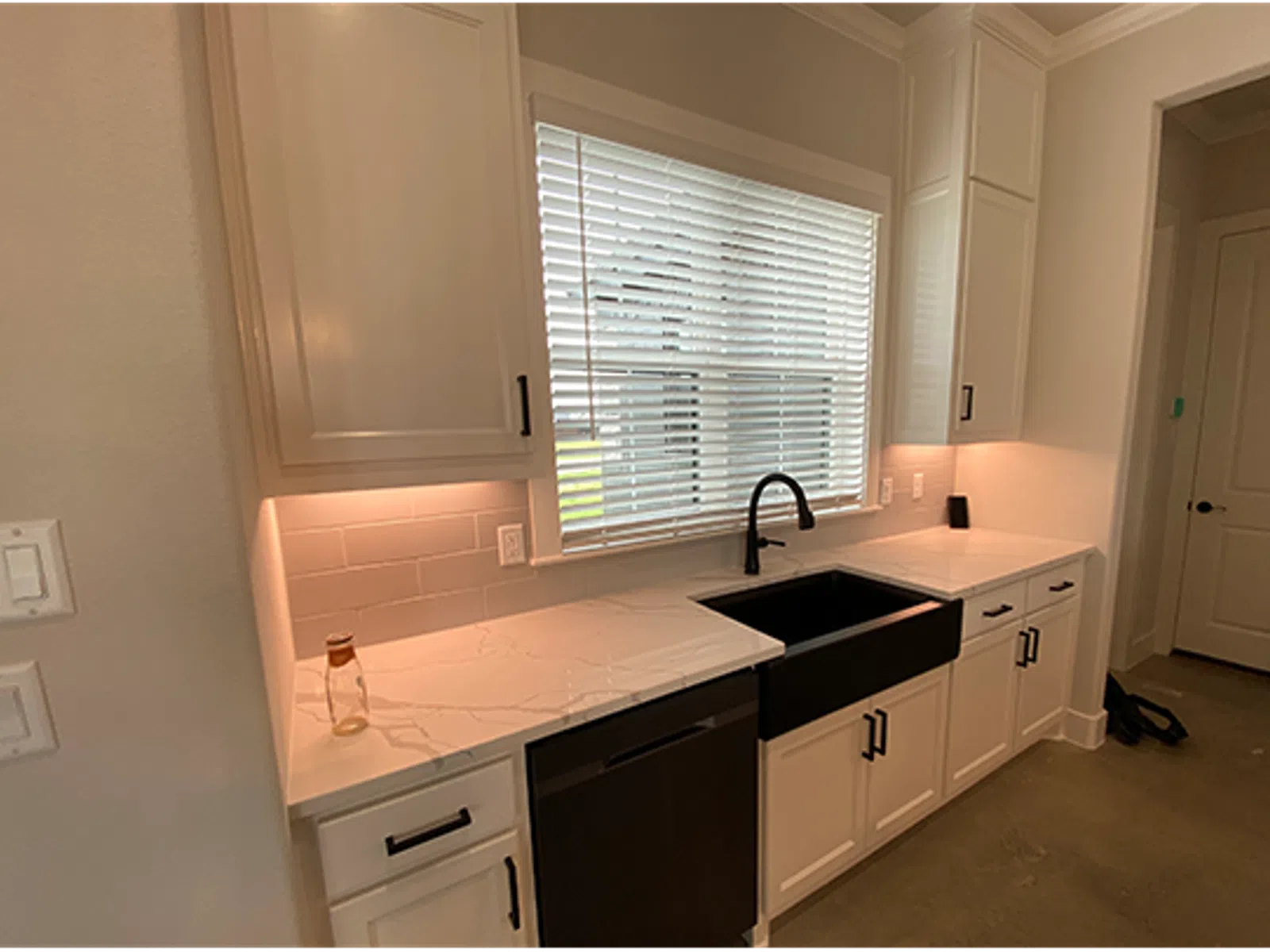 Modern kitchen counter with white cabinets, black farmhouse sink, black faucet, and window with blinds.
