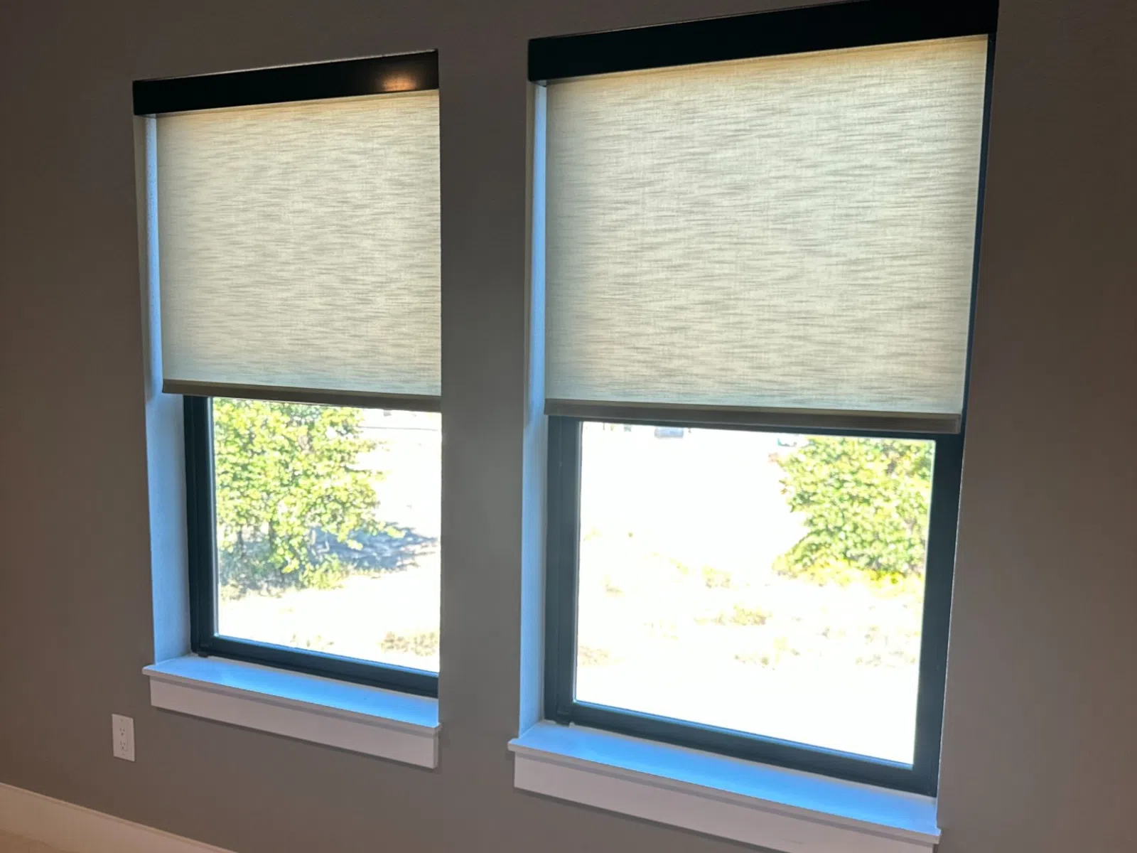Two modern windows with beige roller shades partially drawn in a simple, clean interior room.