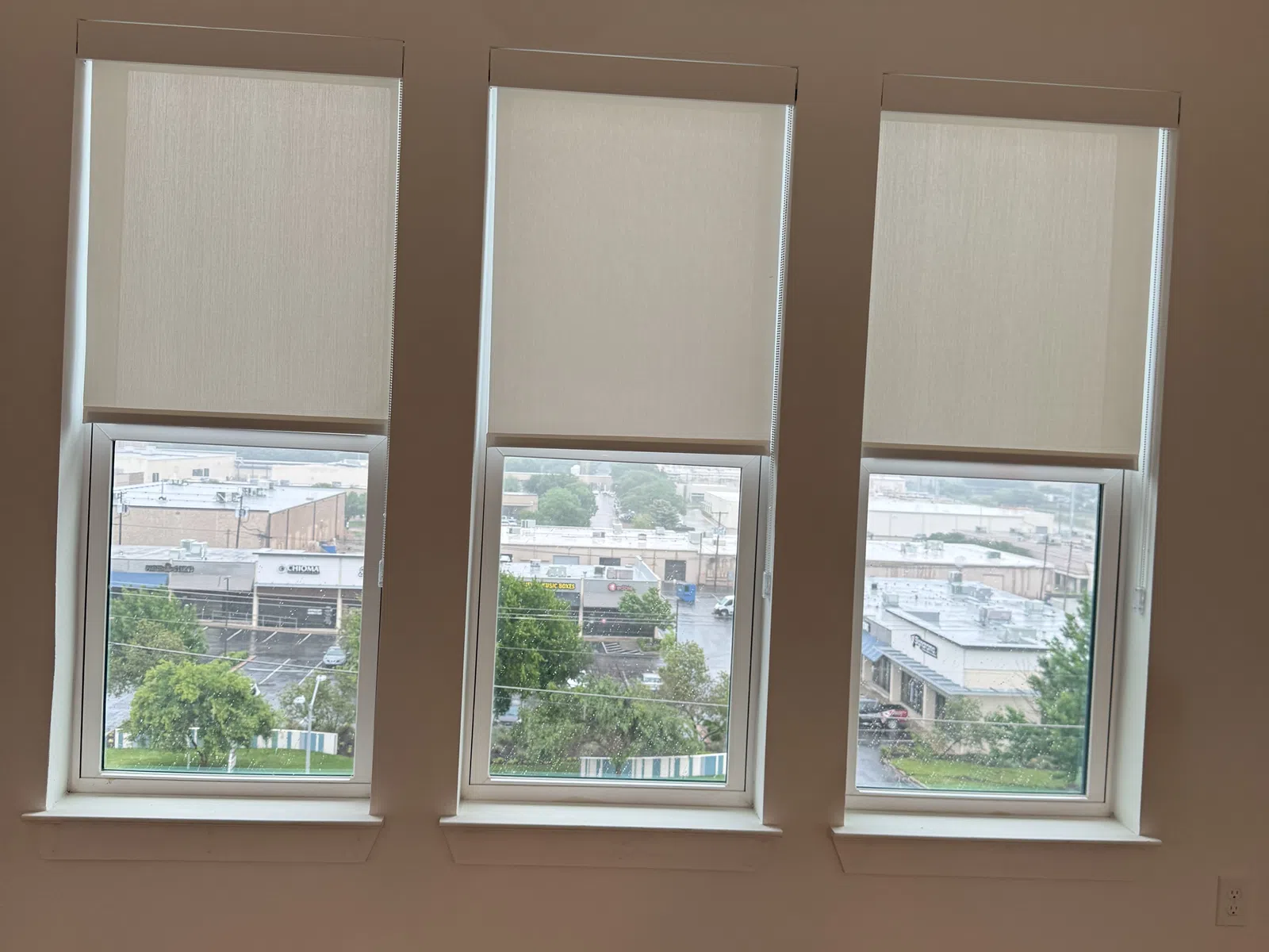 Three tall windows with beige roller blinds partially lowered, showing an urban view outside from a modern room.