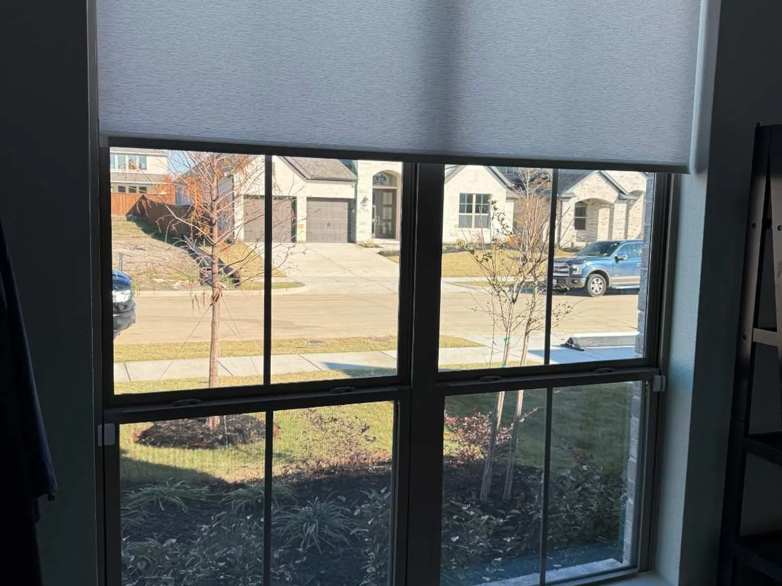 Residential window with white roller shade half lowered, showing a suburban street and houses outside.