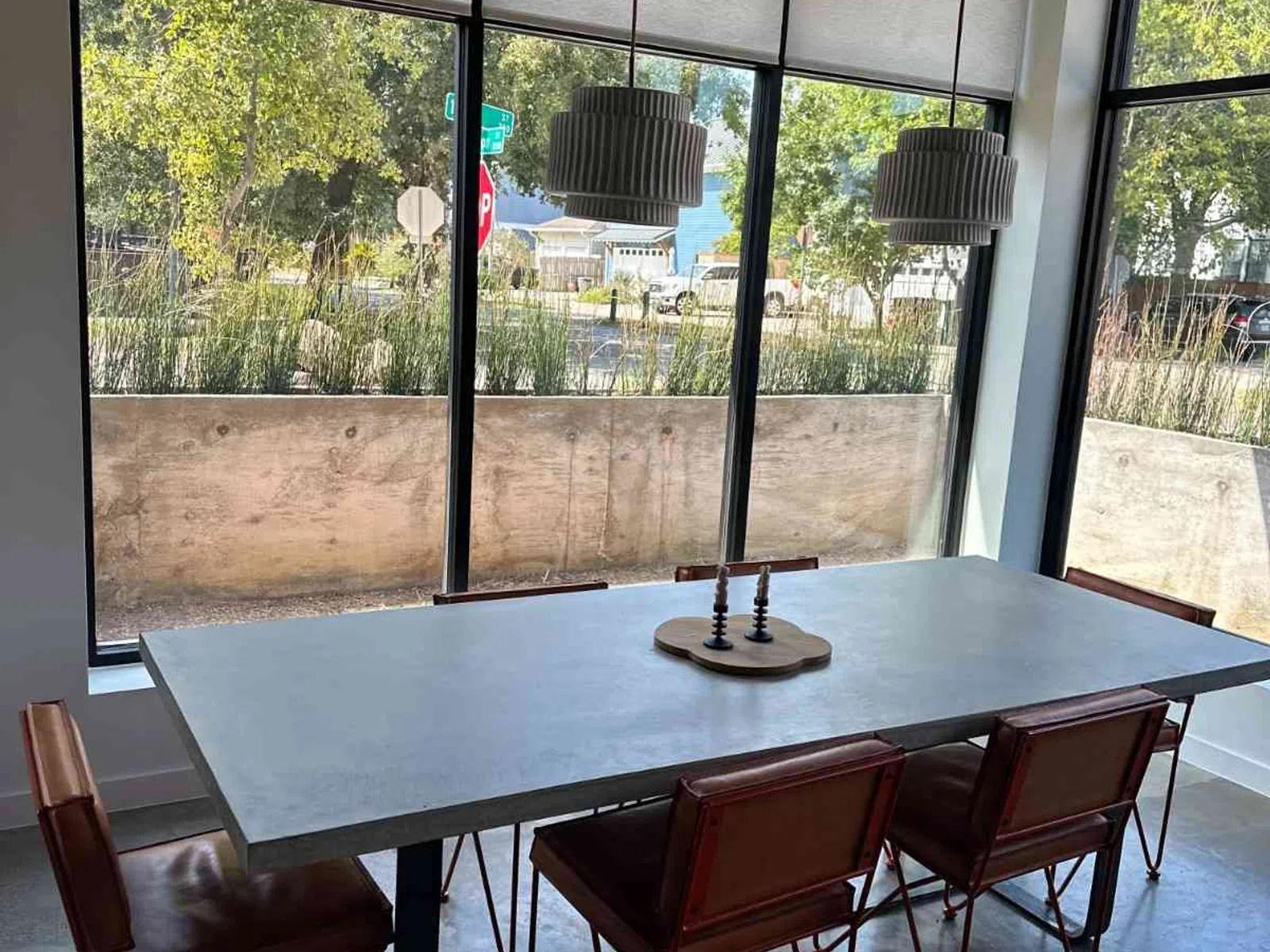 Modern dining room with concrete table, leather chairs, large windows, and pendant lights with outdoor view.