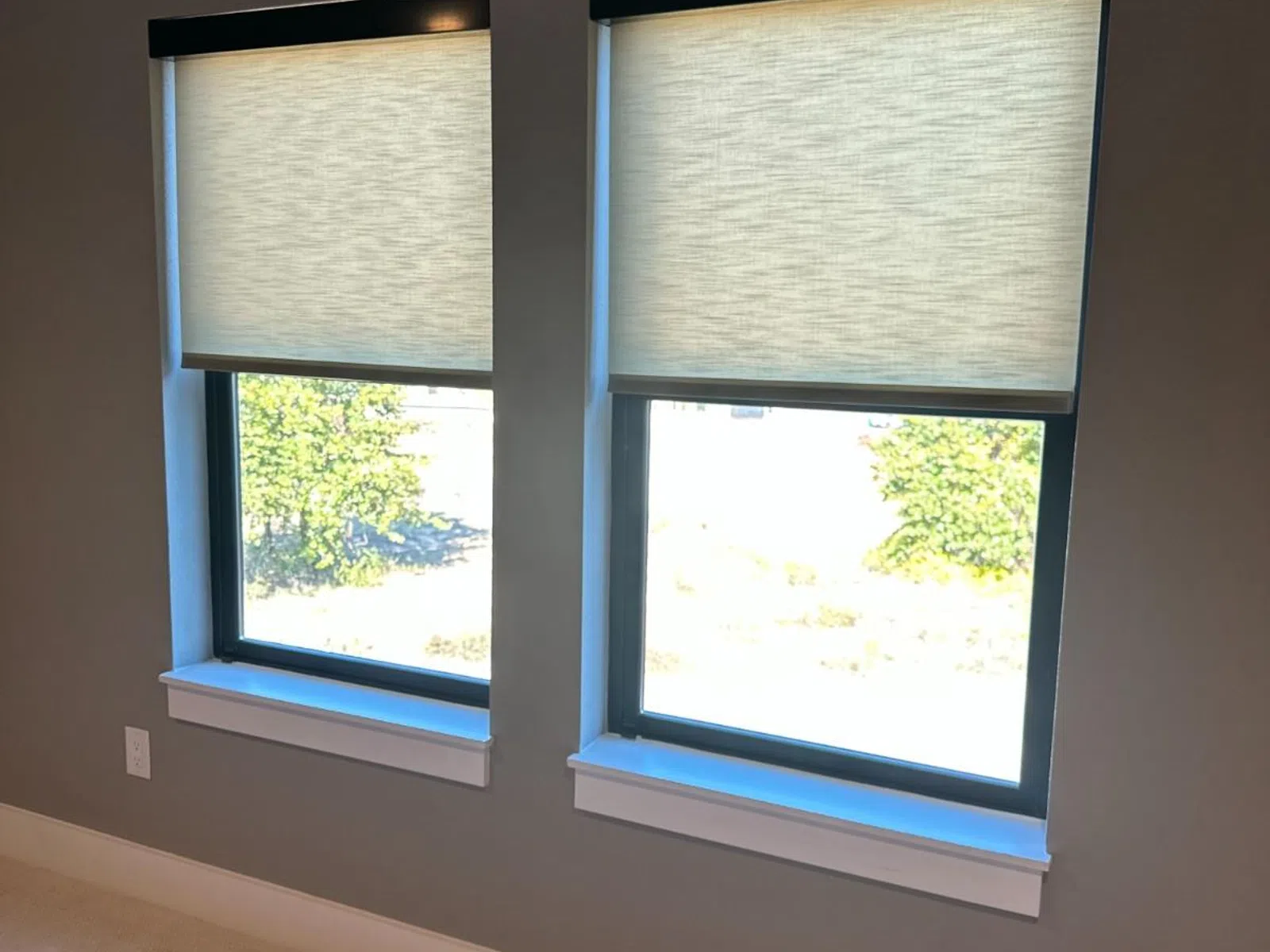 Two windows with light-filtering roller shades partially lowered in a modern room with gray walls.