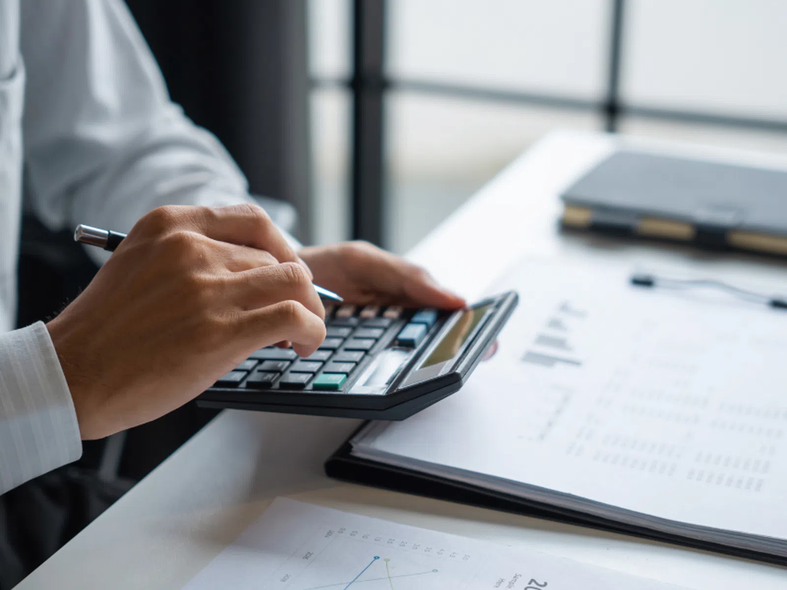 Person using calculator and holding pen over financial documents with charts and graphs on desk.