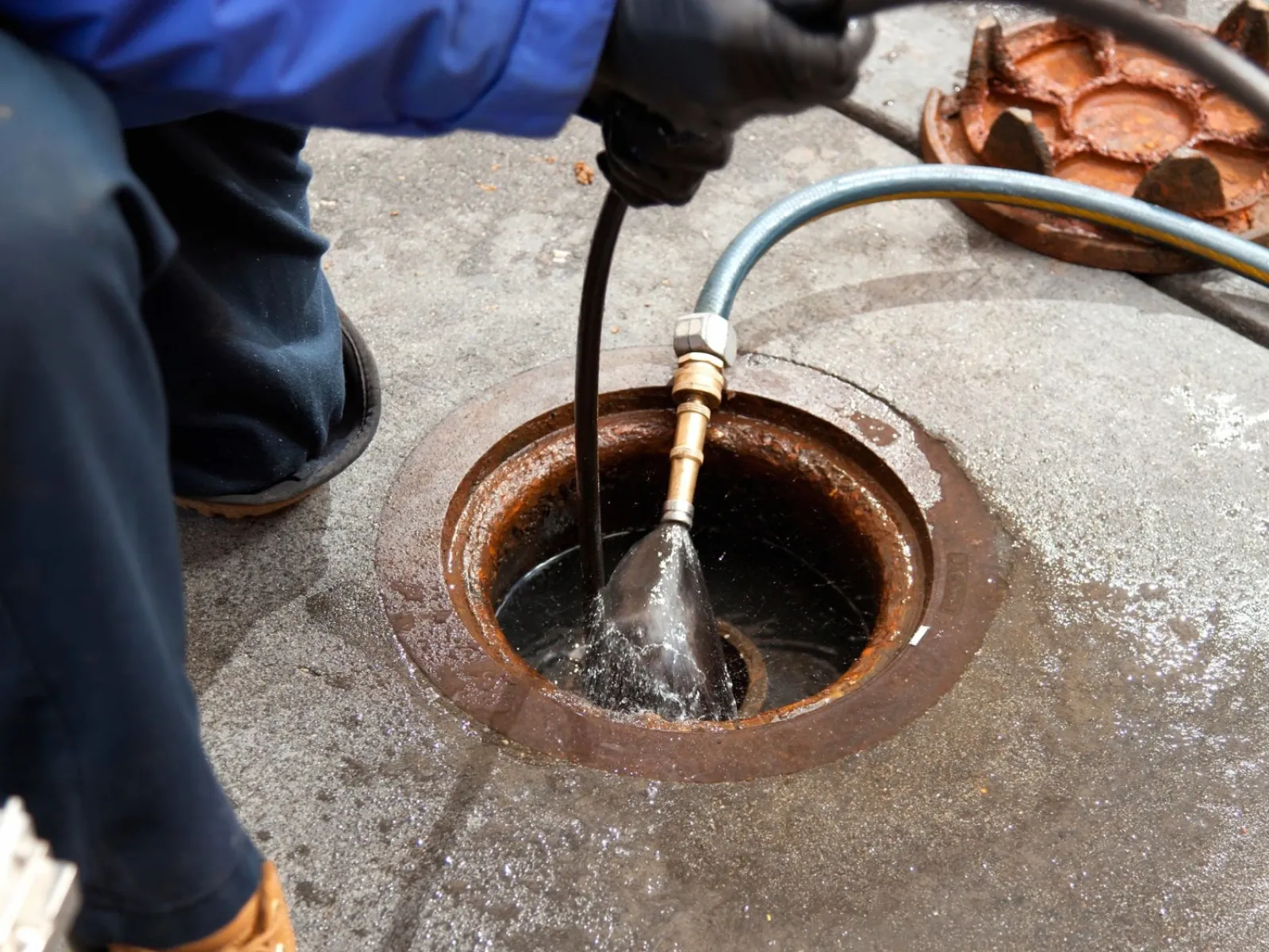 Worker using high-pressure water hose to clean a drain on concrete surface with protective gloves and boots.