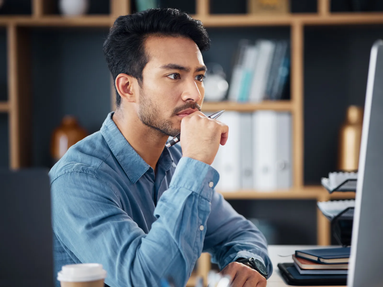 Man in blue shirt thoughtfully working on computer at desk with office shelves in background.