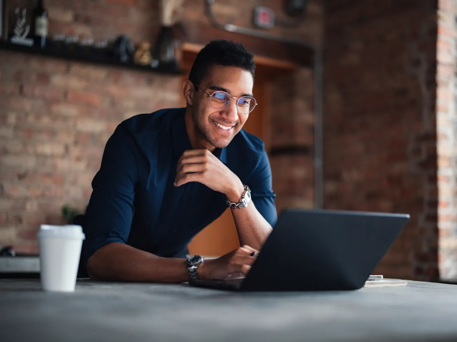 Young man with glasses smiling while working on a laptop in a modern brick-walled office with natural light.