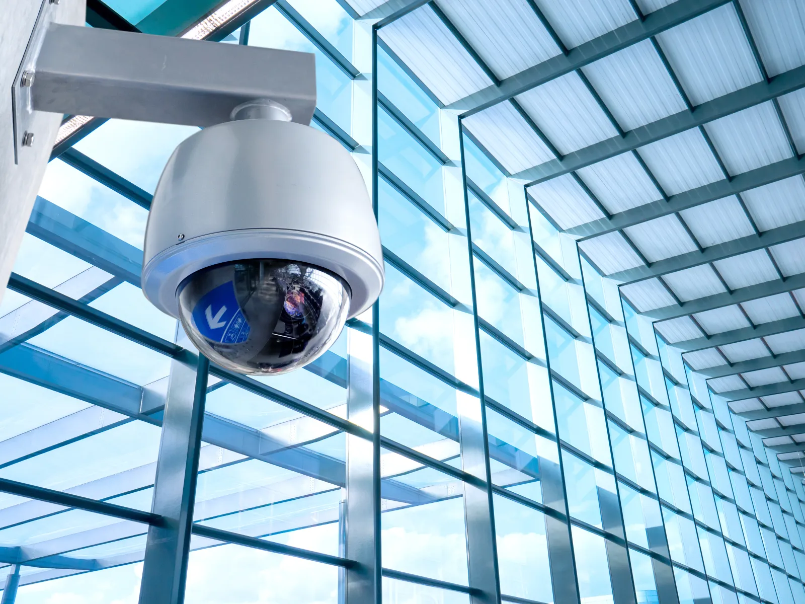 White dome security camera mounted on wall inside modern glass building with blue sky reflection.