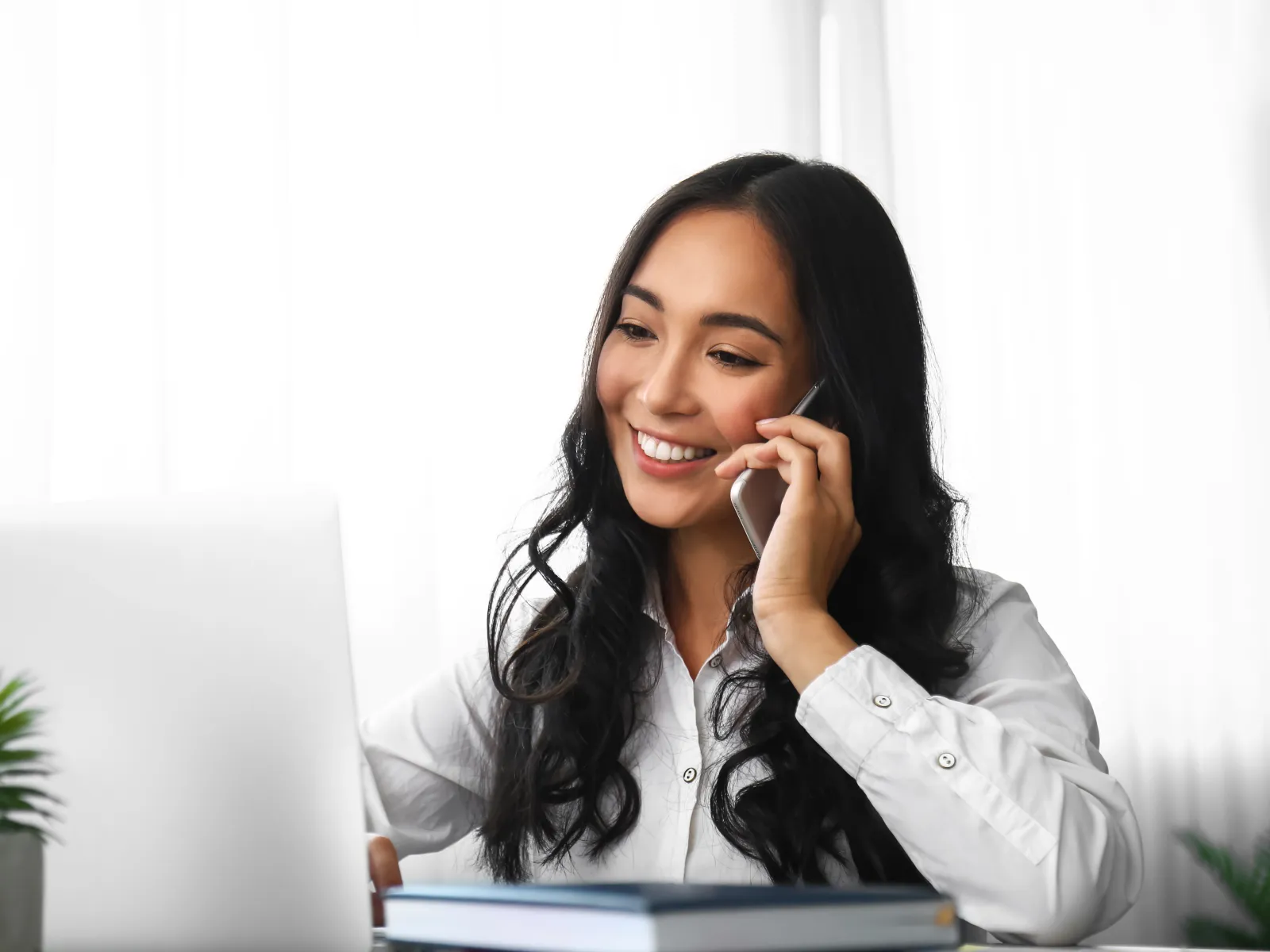 Smiling woman talking on phone while working on laptop at a bright home office desk.