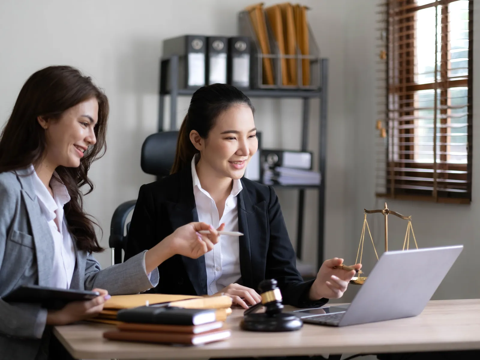 Two female lawyers discussing case files and using a laptop in a modern office with legal symbols present.