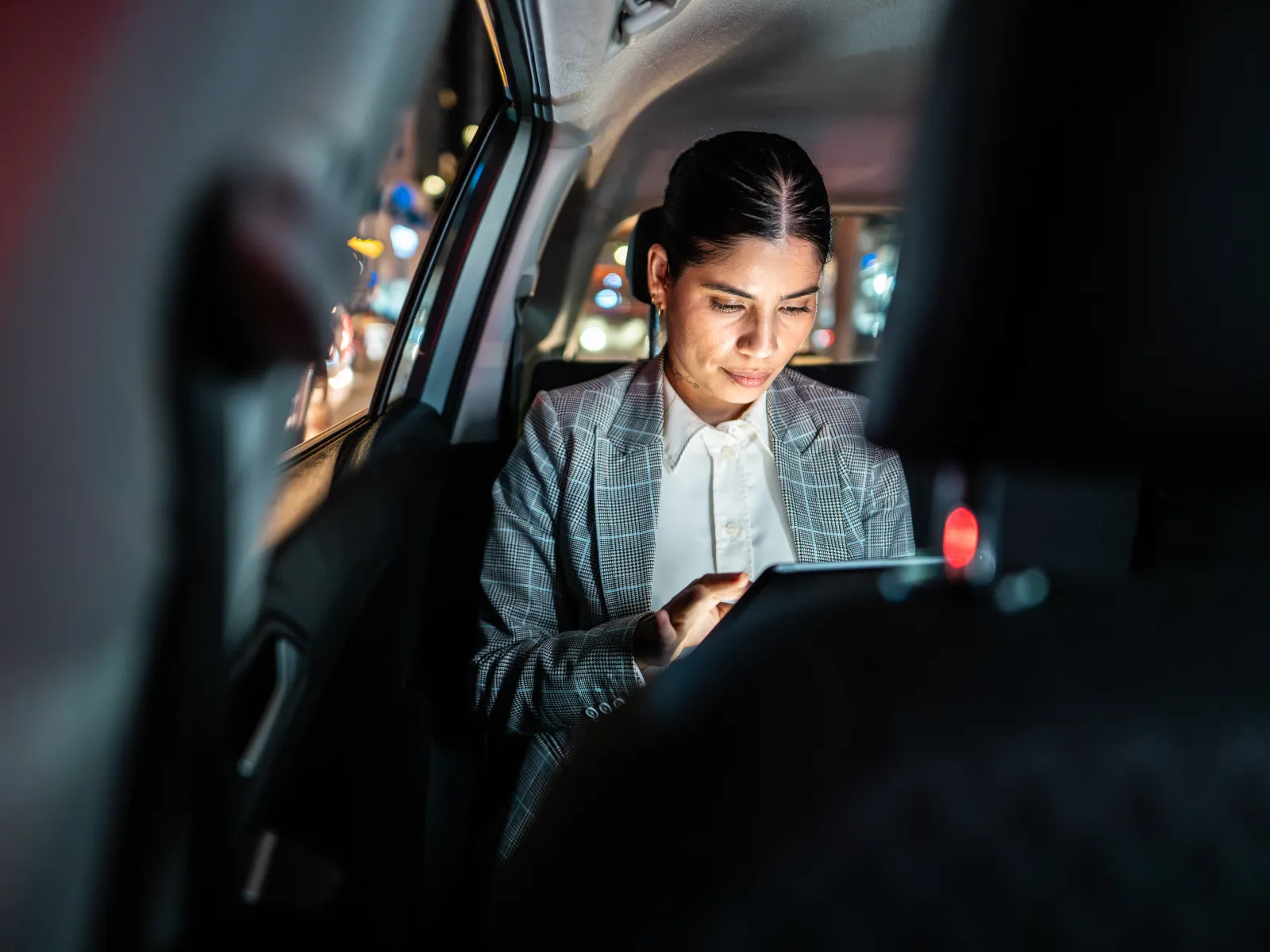 Businesswoman using tablet inside a car at night with city lights visible through the window.