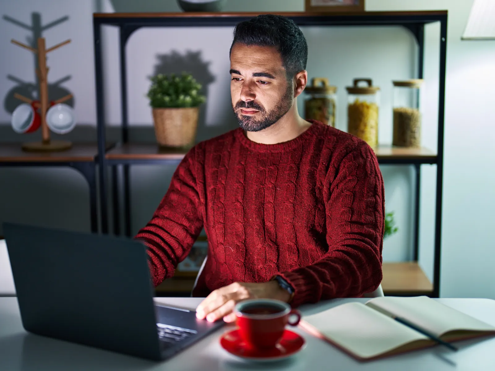 Man in a red sweater working on a laptop at a white table with coffee and an open notebook at home.