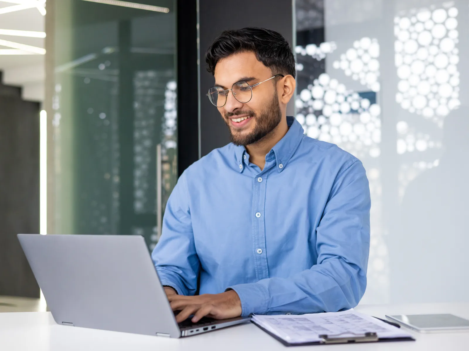 Smiling man in blue shirt working on laptop at modern office desk with clipboard and tablet nearby