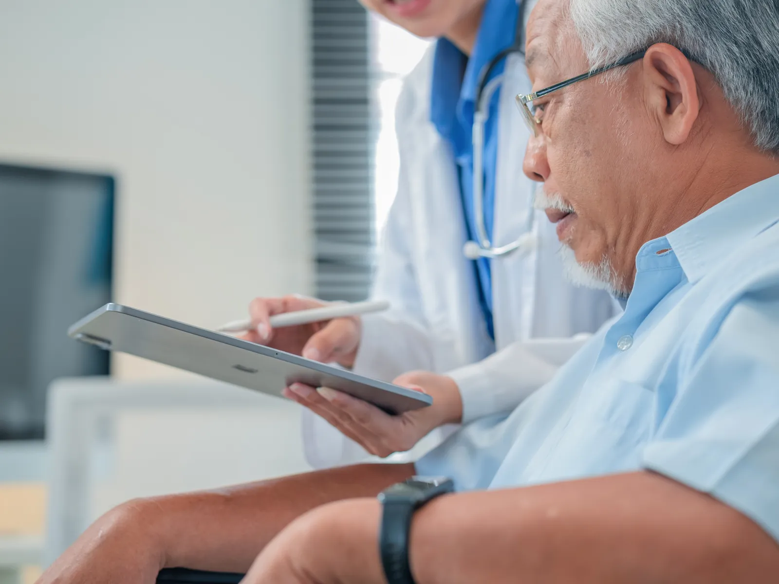 Doctor consulting elderly male patient using a tablet in a clinical setting for medical discussion.