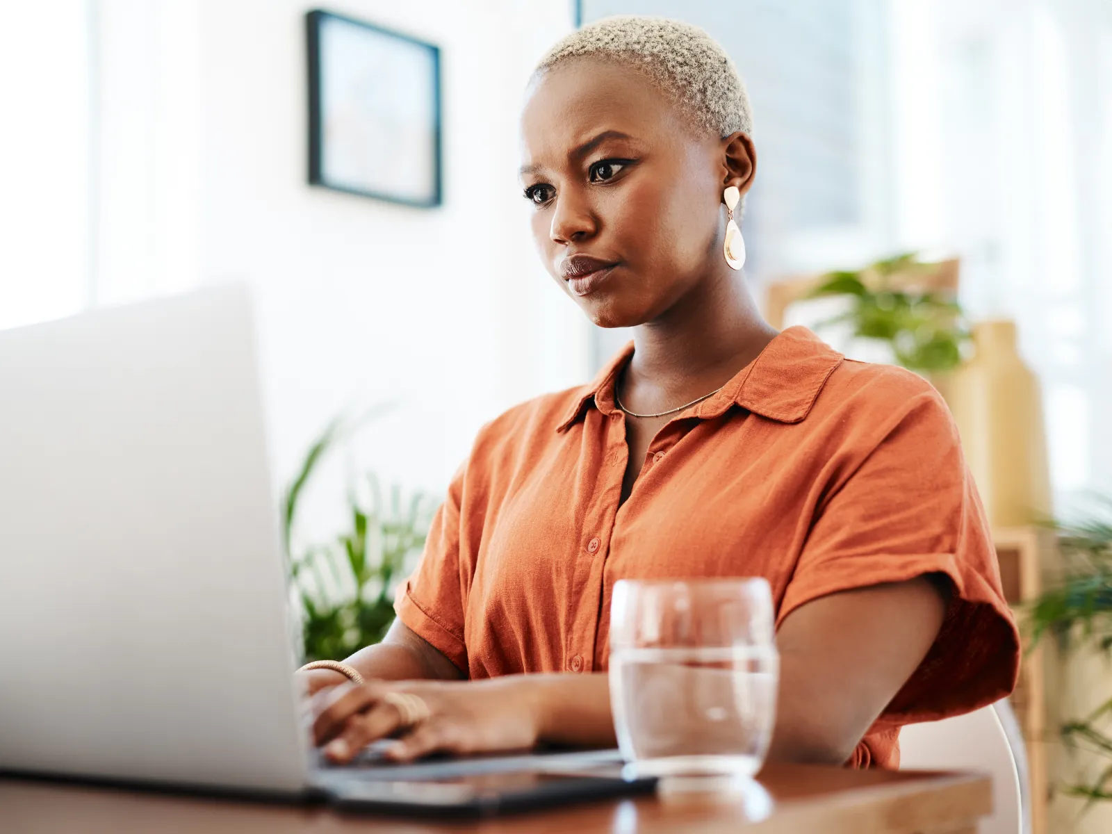 Focused woman in orange shirt working on laptop at a wooden desk with a glass of water nearby in bright room.