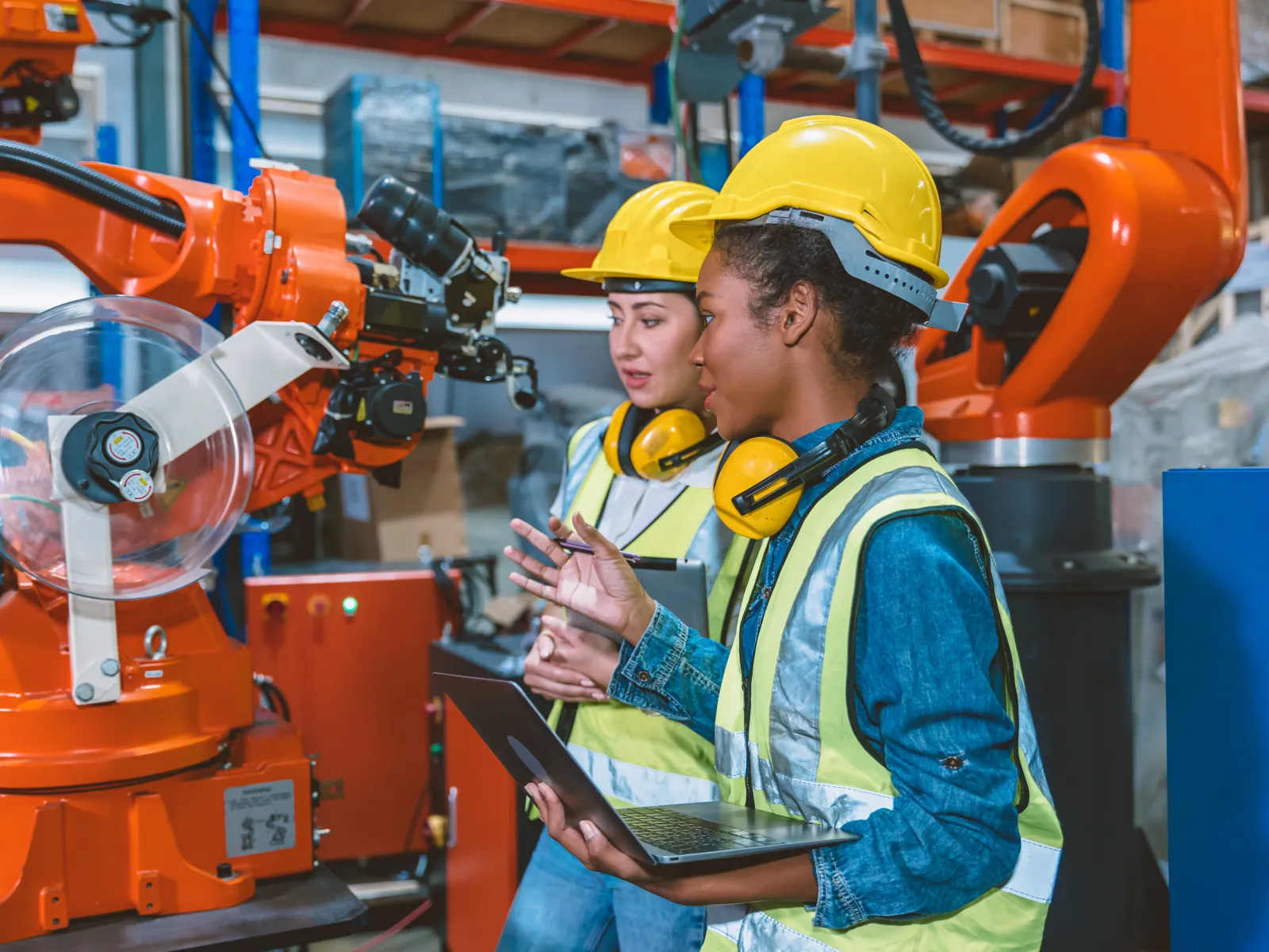 Two female engineers in safety gear inspecting industrial robotic arms in a manufacturing facility.