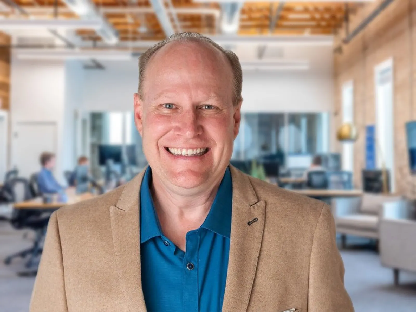 Smiling middle-aged man in blue shirt and tan blazer standing in modern open office with exposed wooden ceiling.