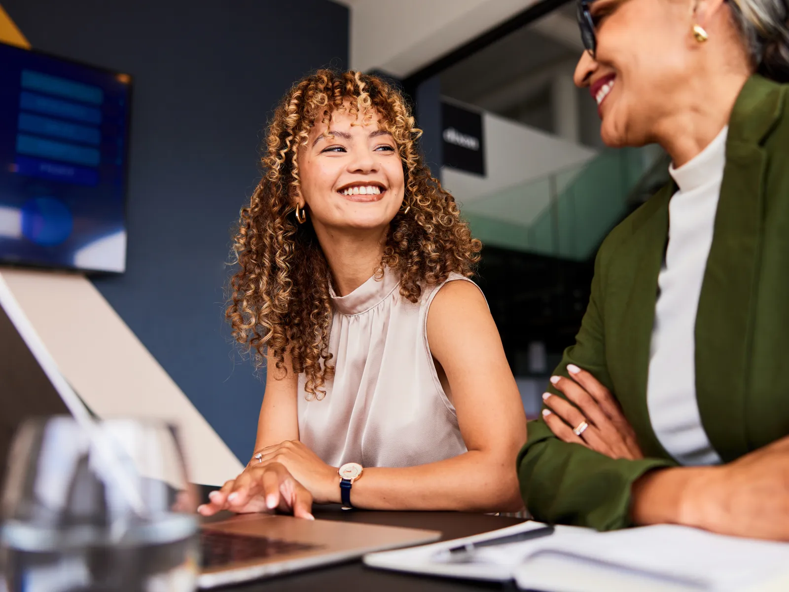 Two professional women smiling and collaborating at a laptop in a modern office setting.