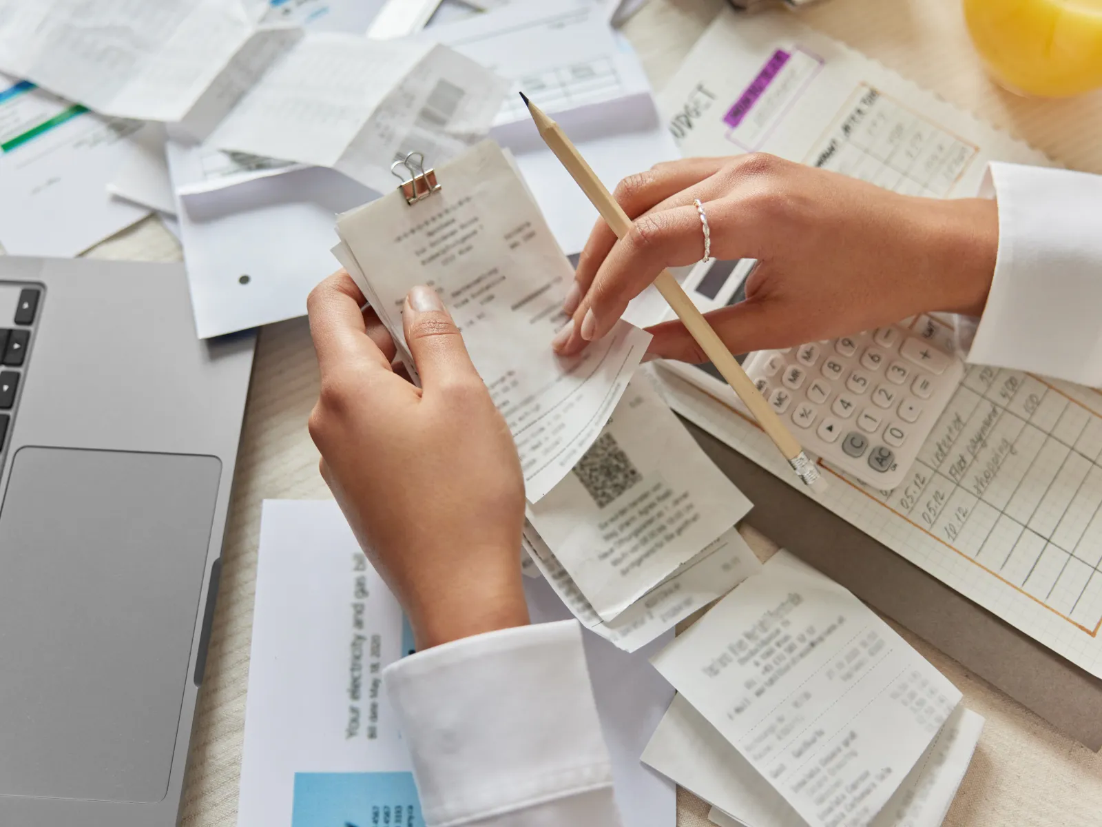 Person sorting receipts and bills with a pencil, calculator, laptop, and documents on a cluttered desk