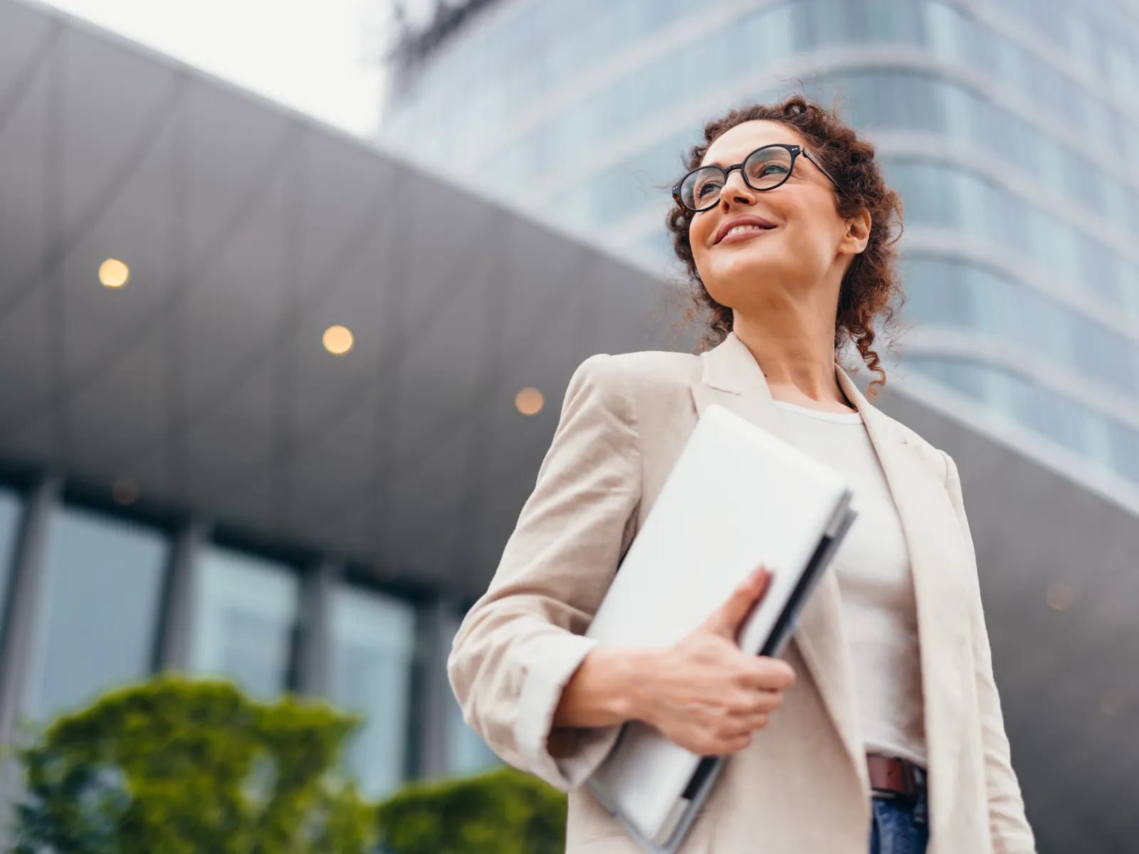 Confident businesswoman in glasses holding laptop outside modern office building smiling upward.