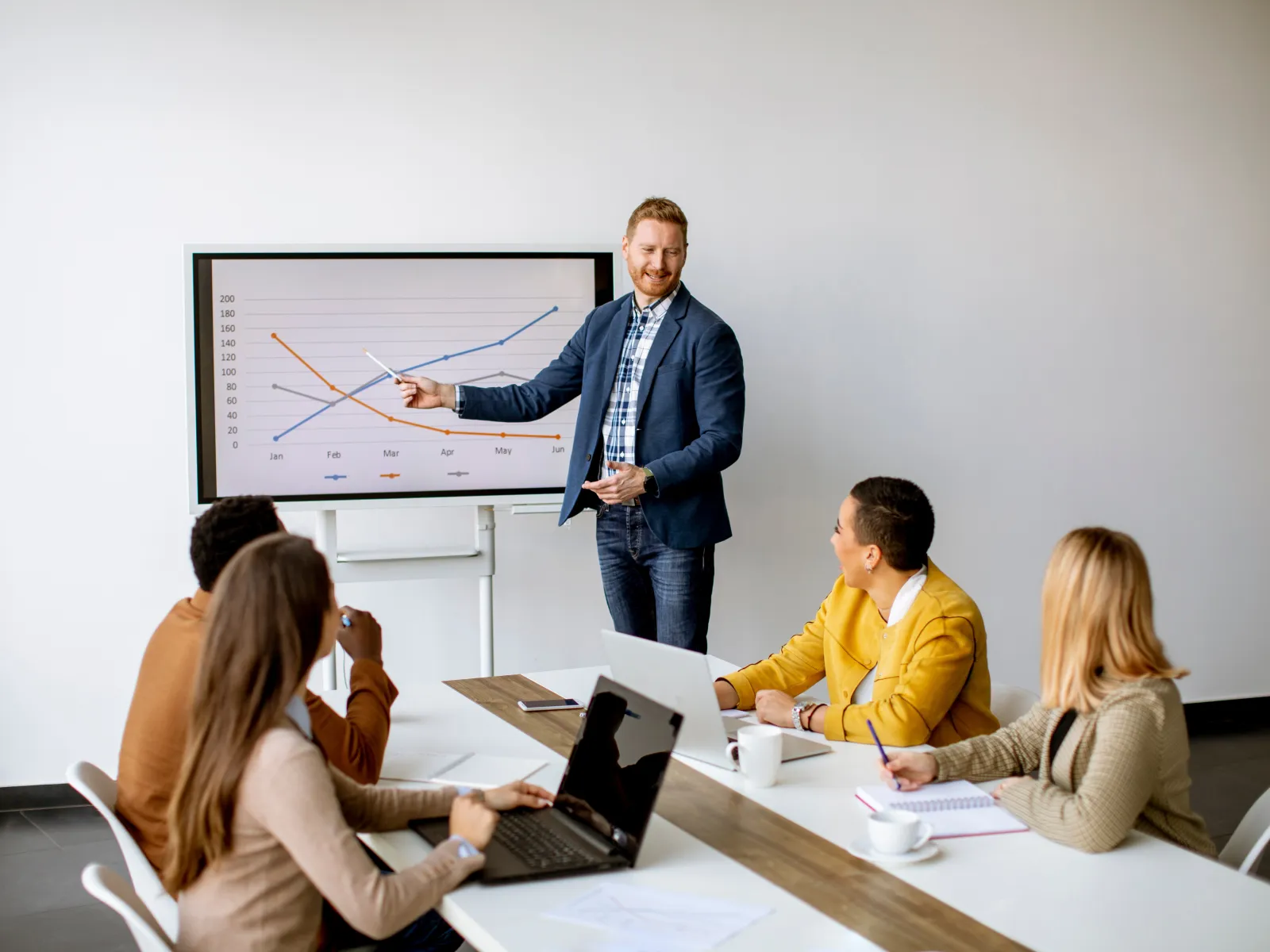 Businessman presenting sales graph to colleagues in a modern meeting room with laptops and notebooks
