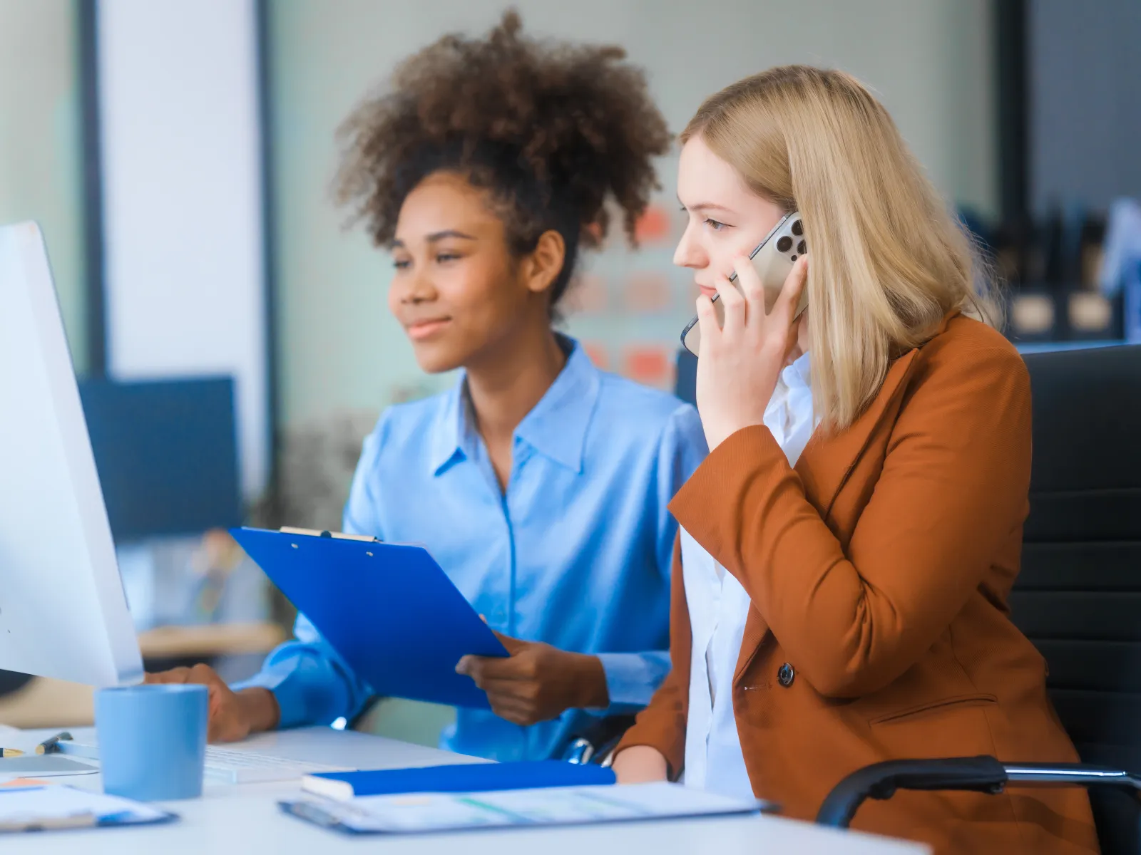 Two professional women collaborate at a desk; one talks on phone, other holds clipboard working on computer.