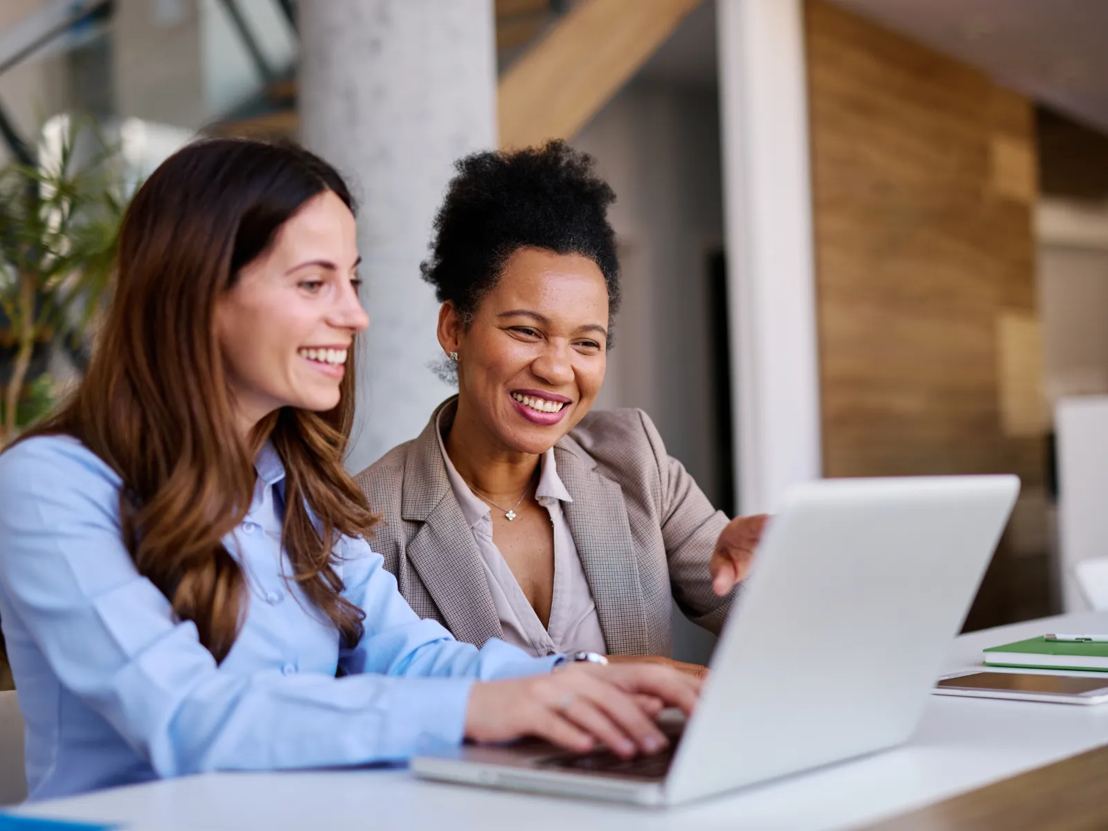 Two women smiling and discussing work while using a laptop in a modern office setting