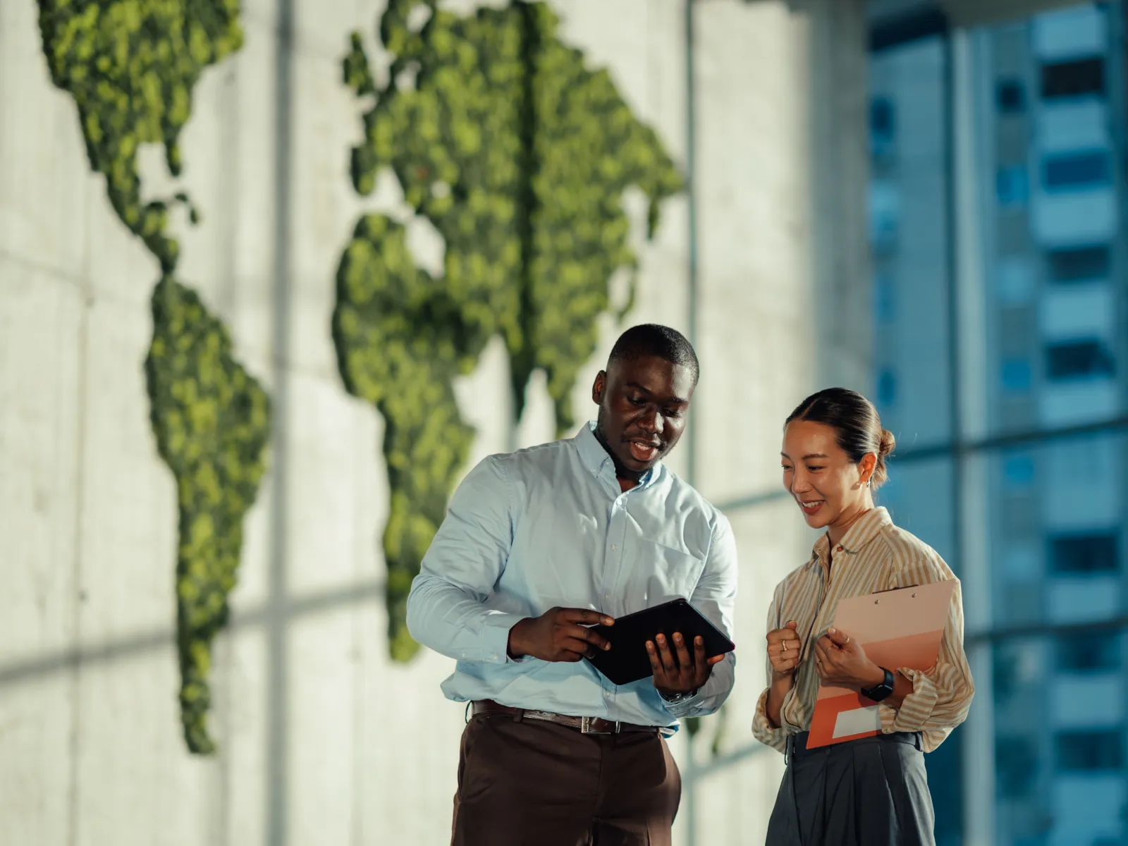 Two professionals discussing work on a tablet in a modern office with a green world map wall art.