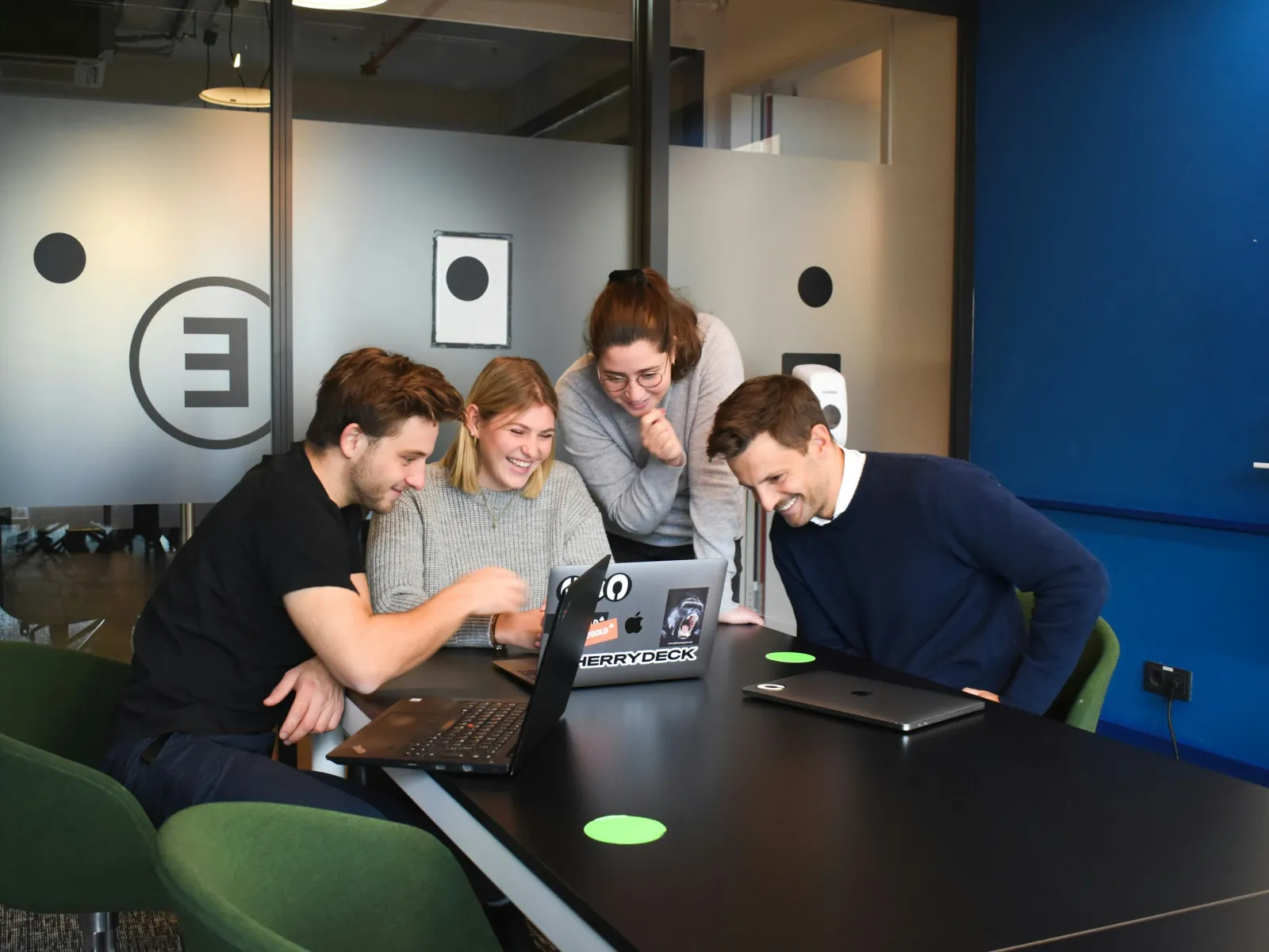 Four young professionals smiling and collaborating around laptops in a modern office meeting room.