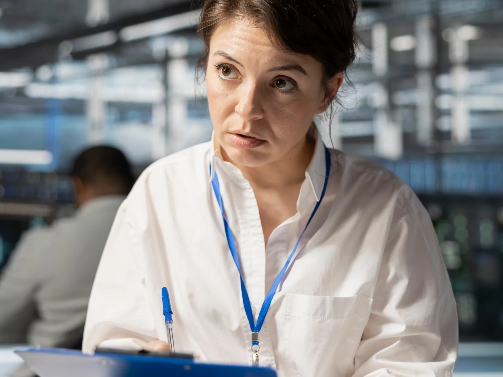Woman in white shirt holding blue clipboard and pen, appearing thoughtful in an industrial workplace.