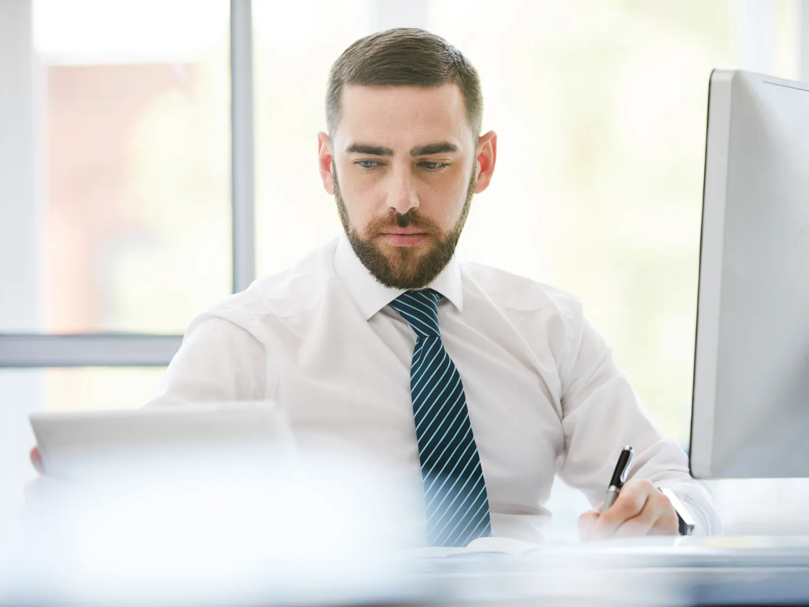 Focused businessman in white shirt and tie working at desk with tablet and computer in bright office.