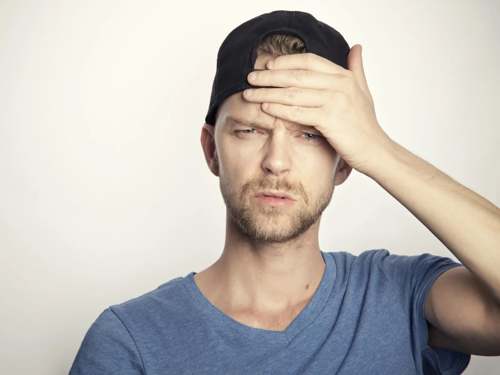 Young man in blue shirt and black cap touching forehead with a concerned expression against plain background