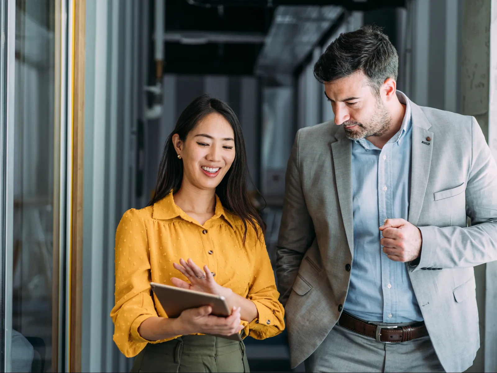 Two professionals discussing work on a tablet in a modern office hallway, smiling and engaged.