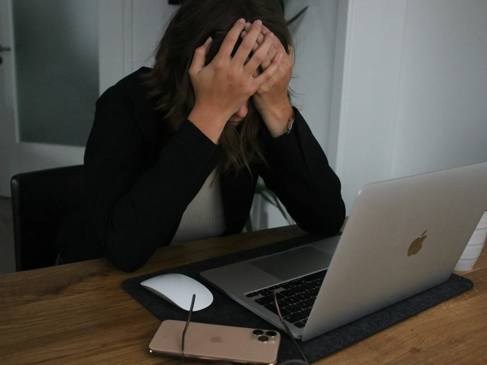 Woman sitting at desk with hands on head appearing stressed while using laptop and smartphone.