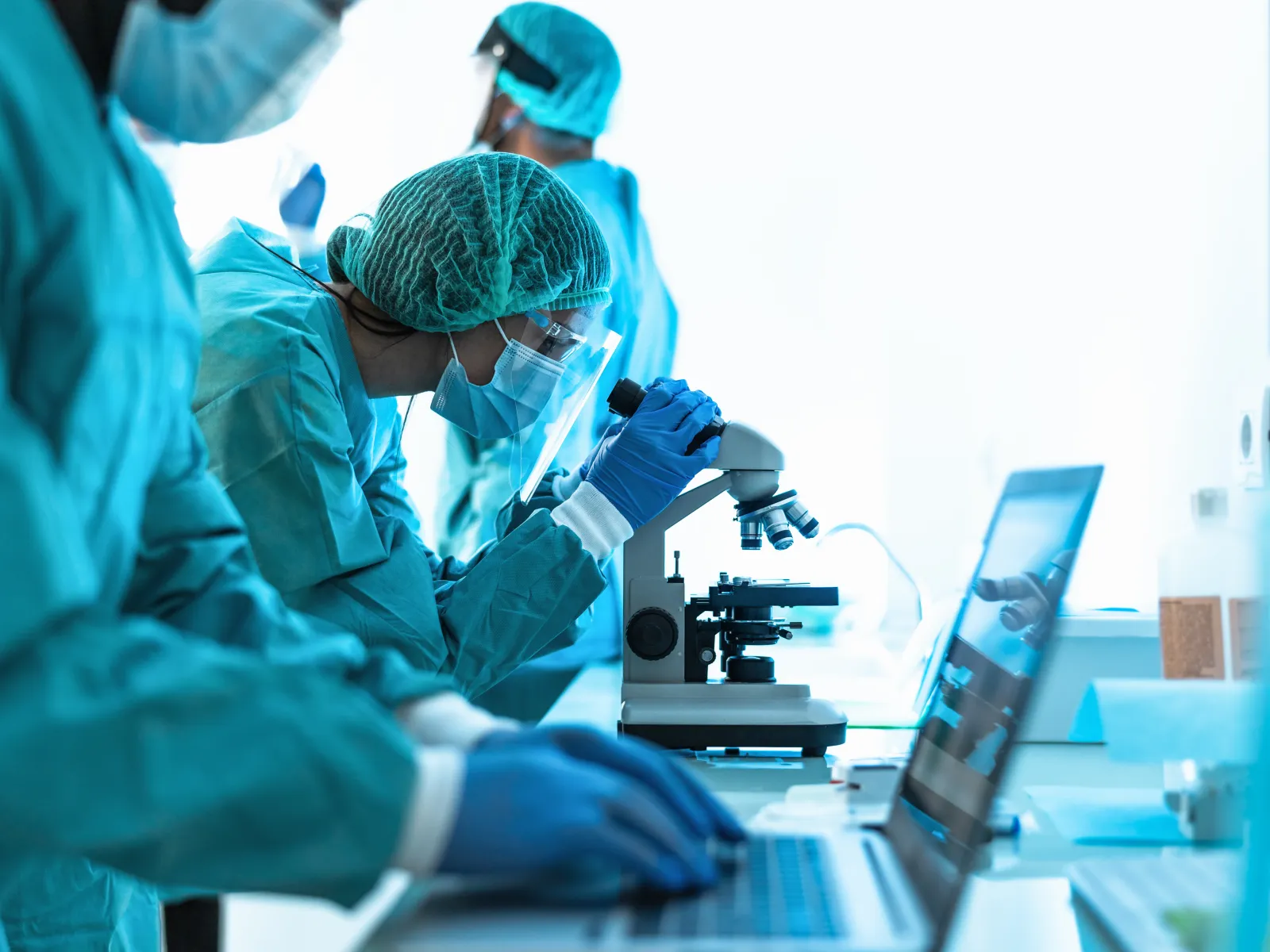 Medical researchers in protective gear using microscope and laptop in a bright laboratory setting.