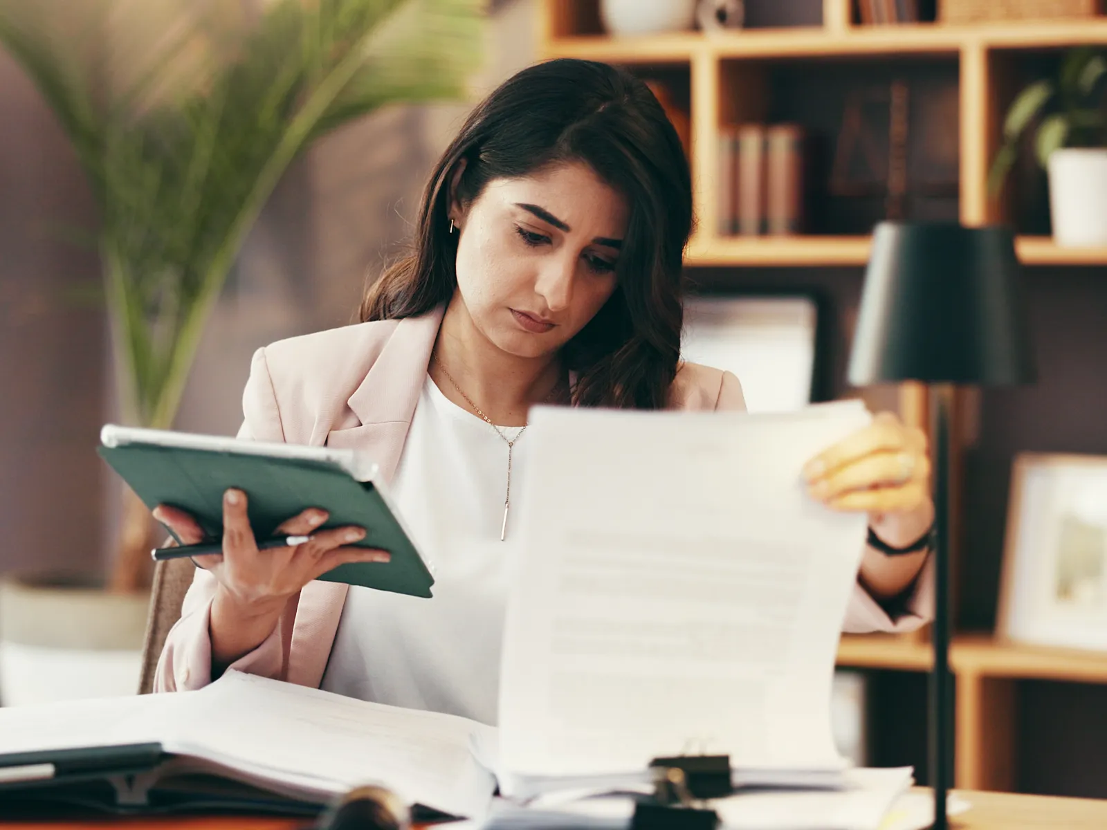 Woman in office reading documents and using a tablet at a desk surrounded by books and plants.