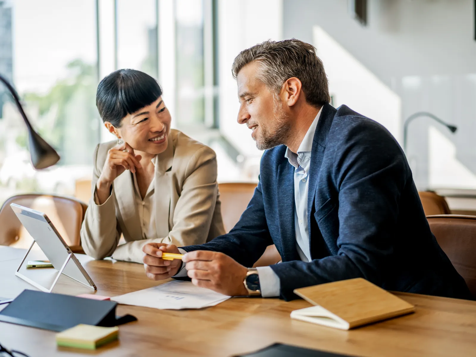 Two professionals engaged in a discussion at a modern office desk with tablet and documents.