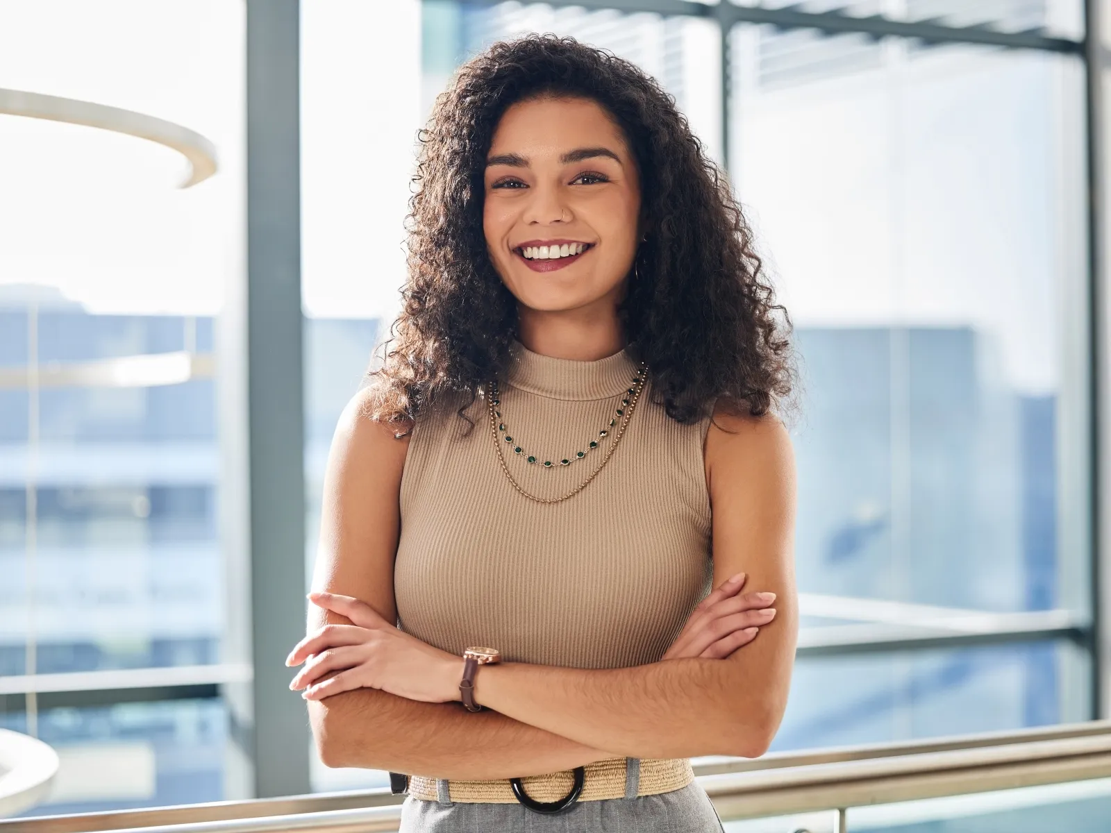 Confident young woman with curly hair wearing beige top and layered necklaces smiling in modern office space