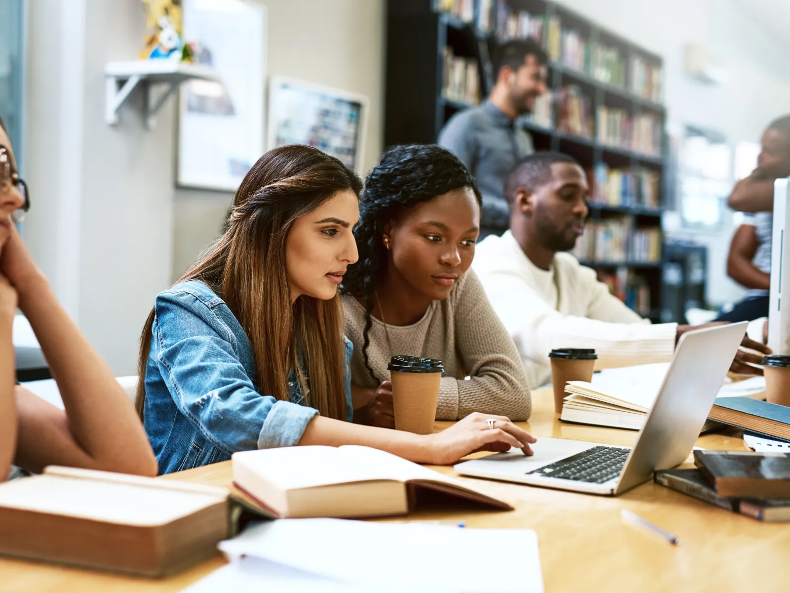 Group of diverse students studying together with laptop and books in a bright library setting.