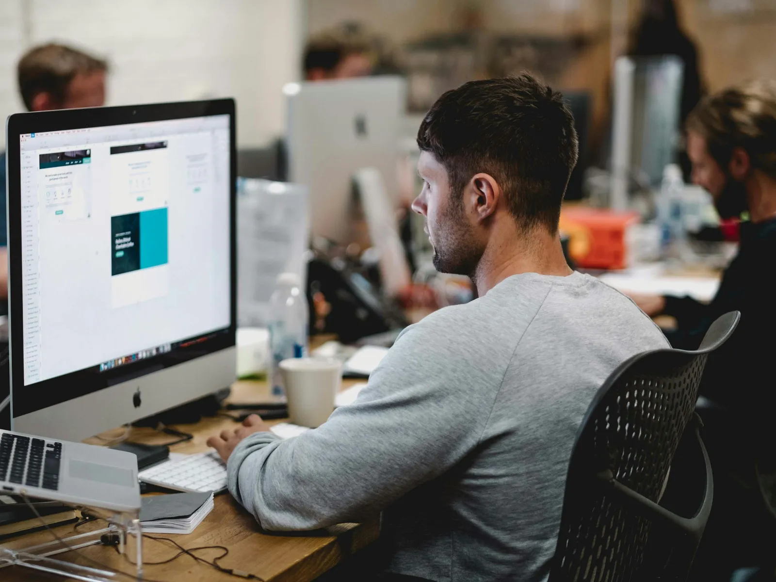 Man working on a desktop computer in a modern office with colleagues and design software on screen
